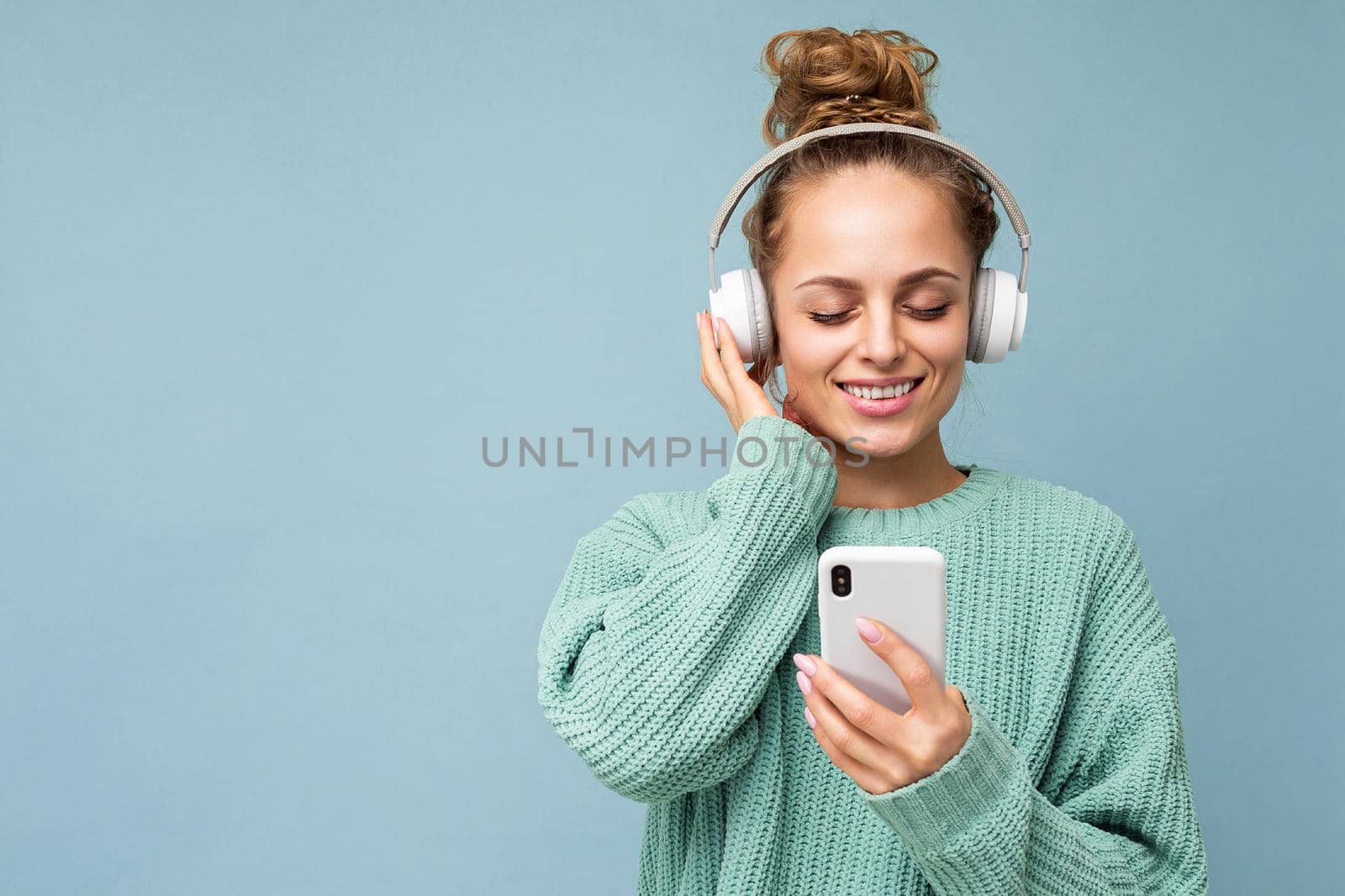 Photo of beautiful joyful smiling young woman wearing stylish casual clothes isolated over background wall holding and using mobile phone wearing white bluetooth headphones listening to music and having fun by TRMK