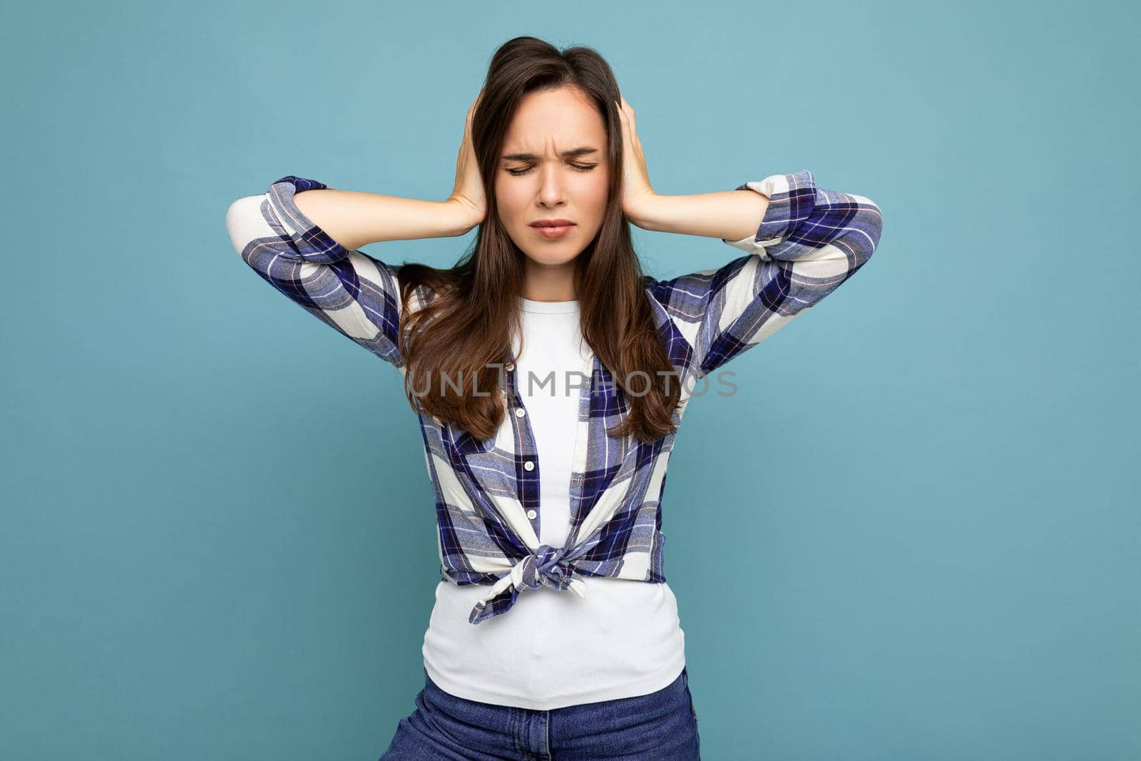 Don't want to hear it. Young emotional beautiful brunette woman wearing check shirt isolated on blue background with copy space and covering ears.