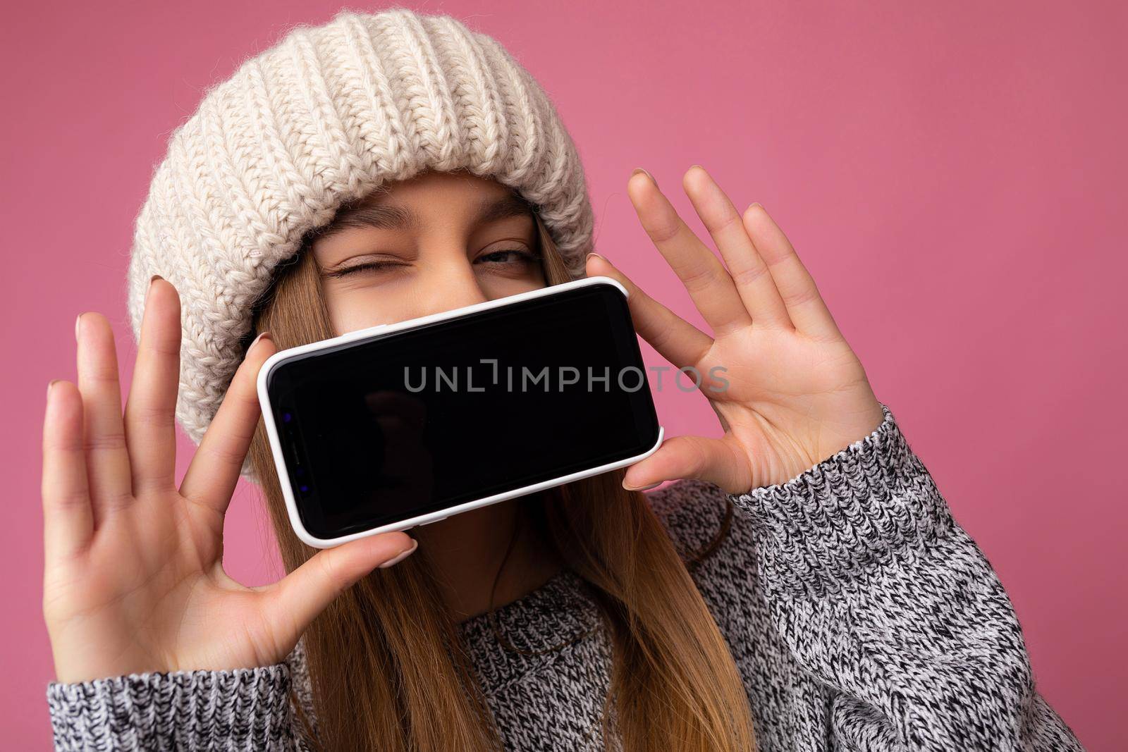 Closeup photo of winking sexy Beautiful young blonde woman wearing casual grey sweater and beige hat isolated over pink background holding in hand and showing mobile phone with empty display for mockup looking at camera by TRMK