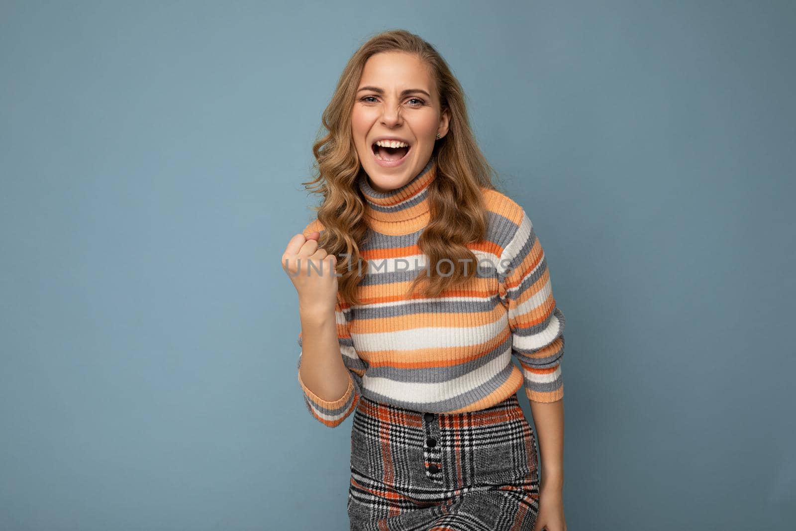 Portrait of happy positive young attractive blonde curly woman with sincere emotions wearing trendy striped pullover isolated on blue background with copy space and celebrating winning.