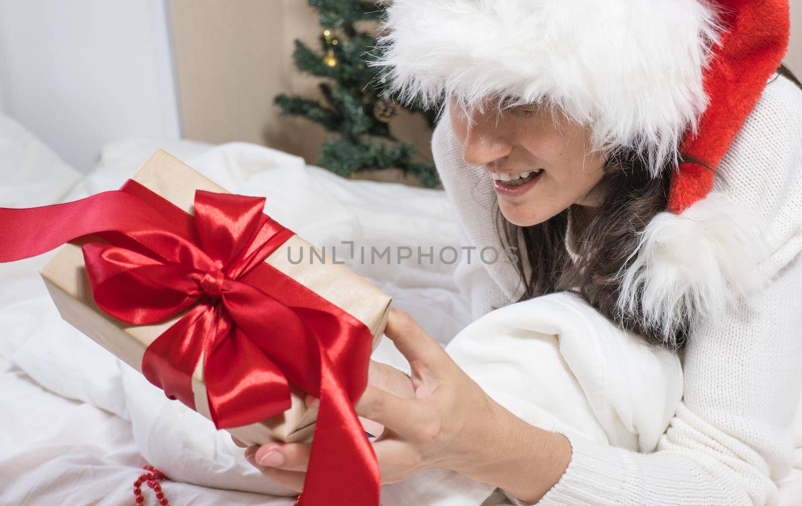 Happy excited young woman in santa claus hat with gift box on white bed by Ekaterina34