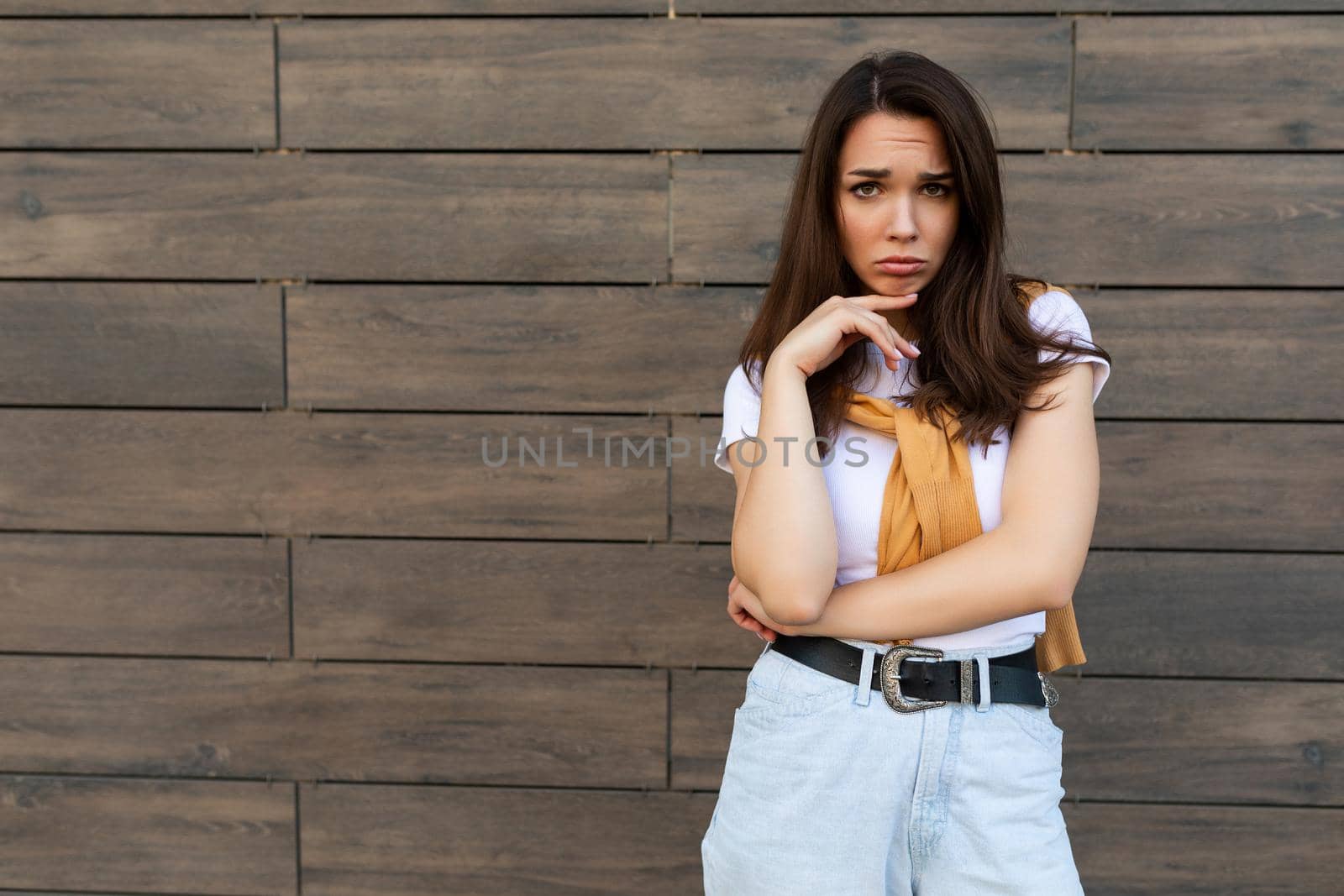 Portrait of touchy sad upset offended young brunet woman wearing casual white t-shirt and jeans with yellow sweater poising near brown wall in the street.