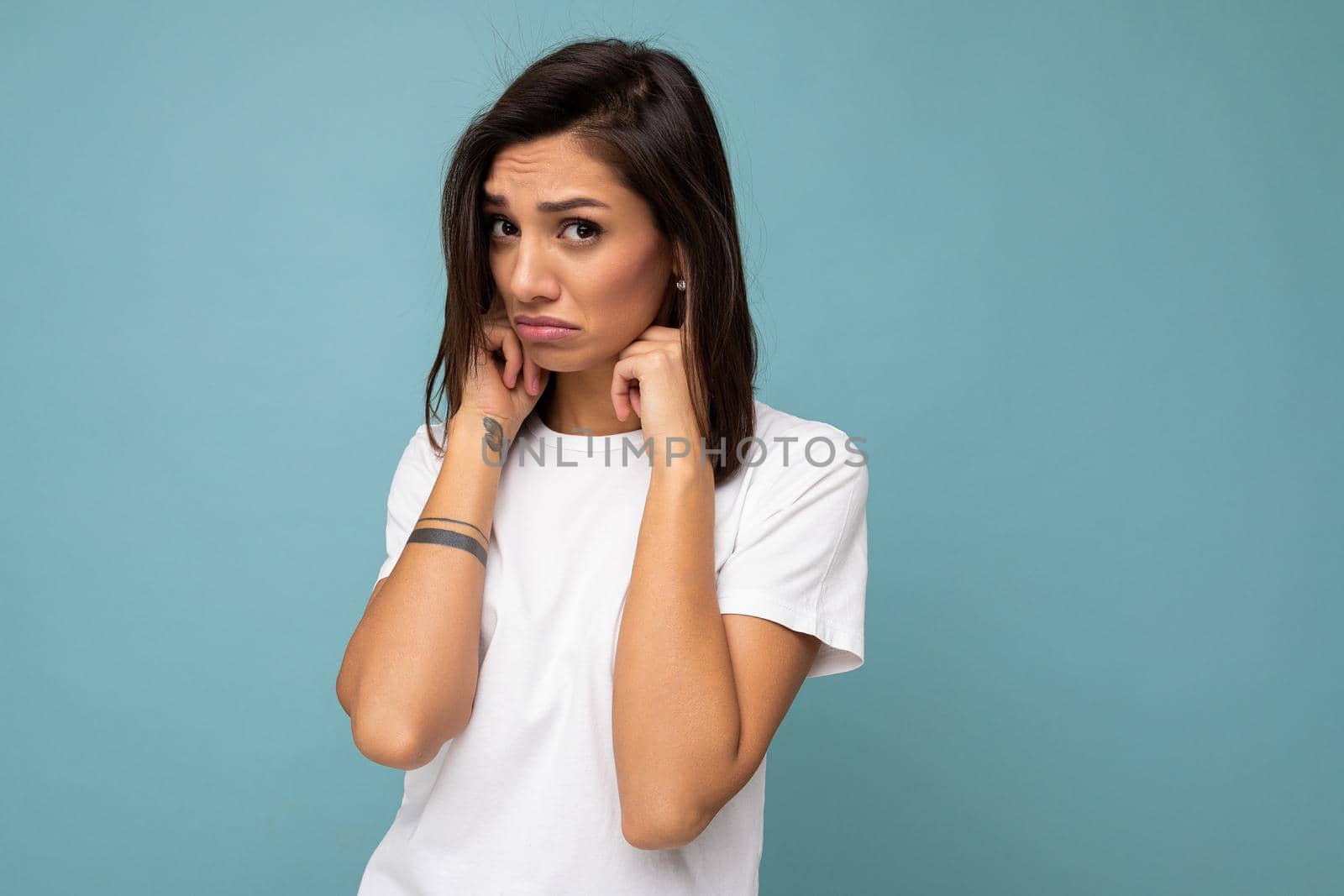 Portrait of sorrowful offended sad young pretty nice brunette woman with sincere emotions wearing casual white t-shirt for mockup isolated on blue background with copy space and covering ears with fingers.