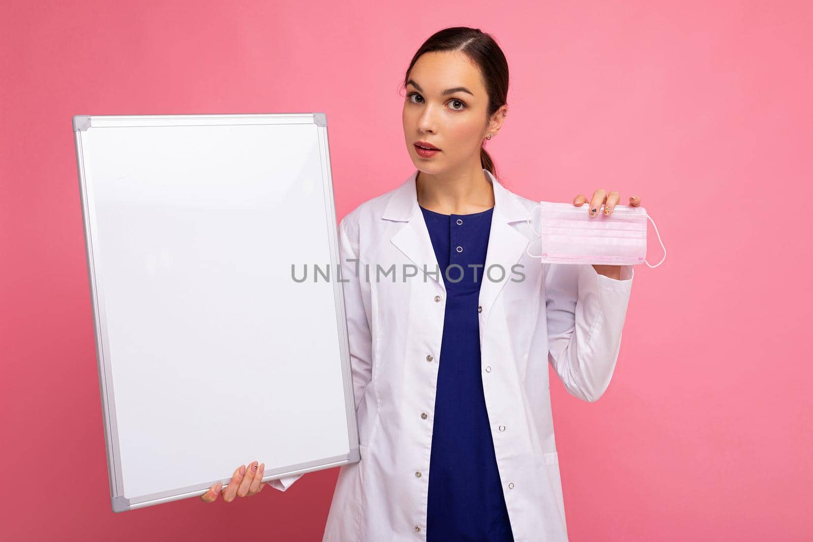 Female person doctor in a white medical coat holding blank board with copy space for text and protective mask isolated on pink background. Epidemic concept.