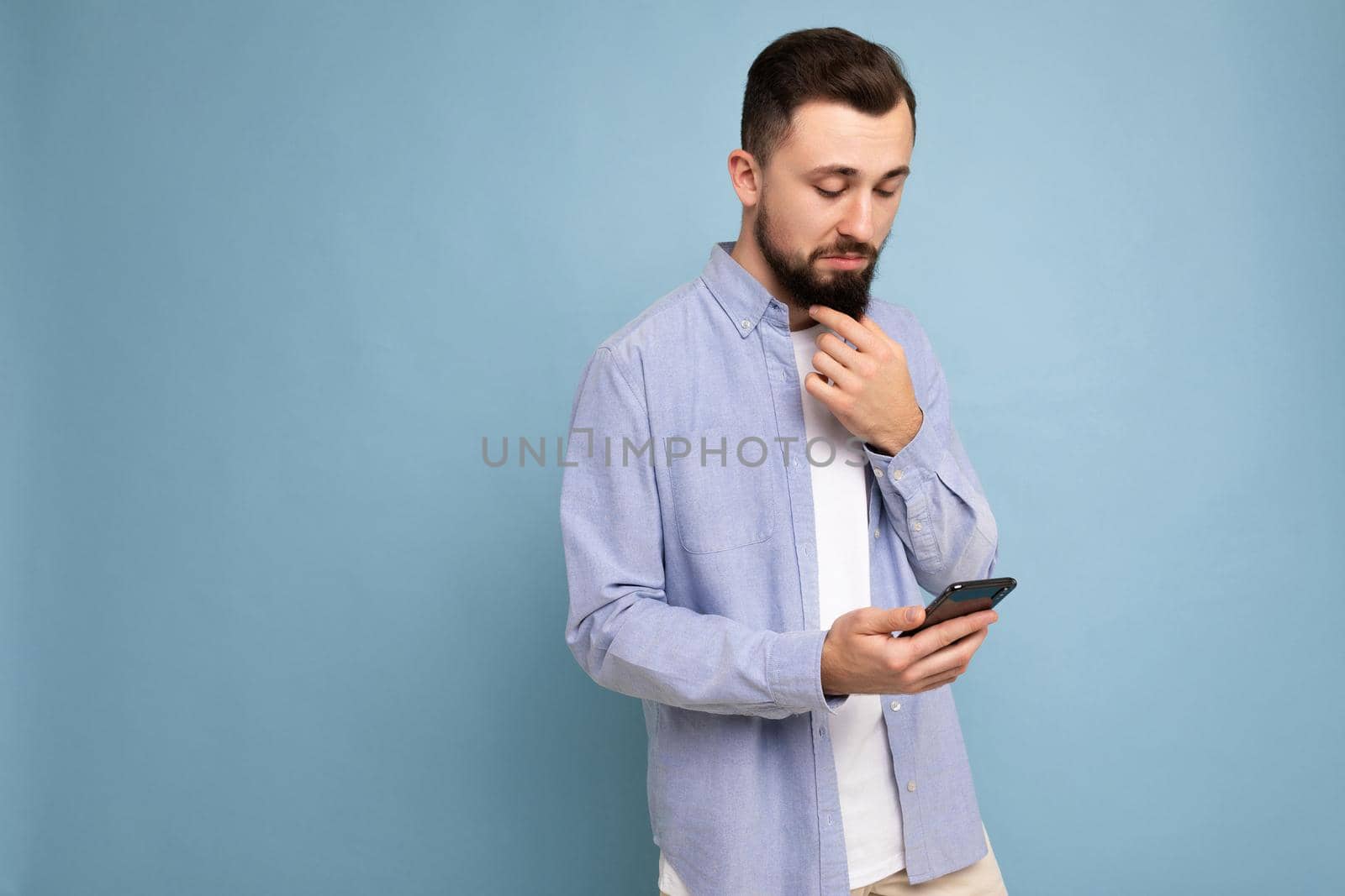 Photo shot of handsome concentrated good looking young man wearing casual stylish outfit poising isolated on background with empty space holding in hand and using mobile phone messaging sms looking at smartphone display screen.