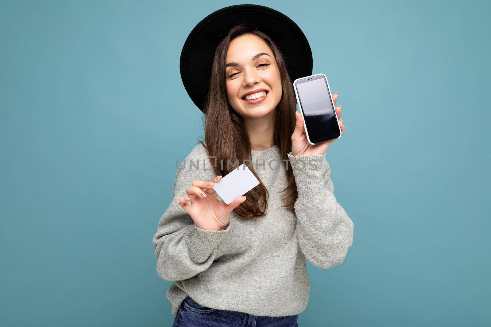 Beautiful positive smiling young brunette woman wearing black hat and grey sweater isolated over blue background holding credit card and mobile phone with empty display for mockup looking at camera by TRMK