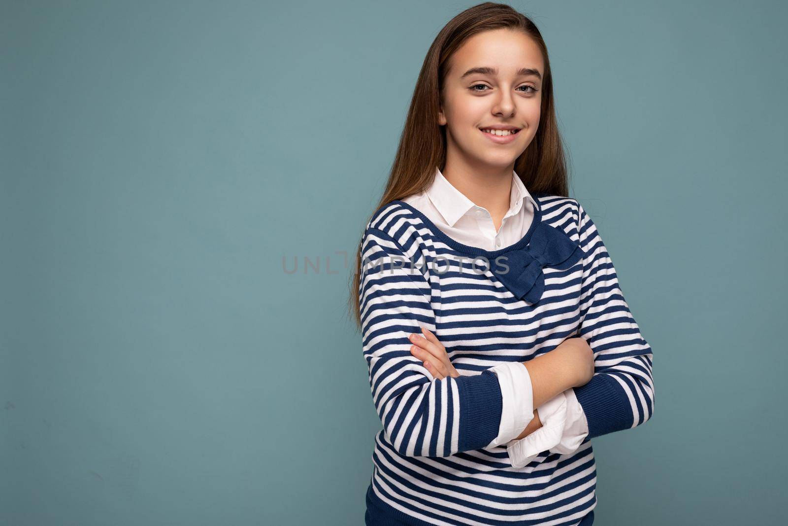 Shot of beautiful positive smiling brunette little girl wearing striped longsleeve standing isolated over blue background wall crossing hands and looking at camera. Empty space, copy space