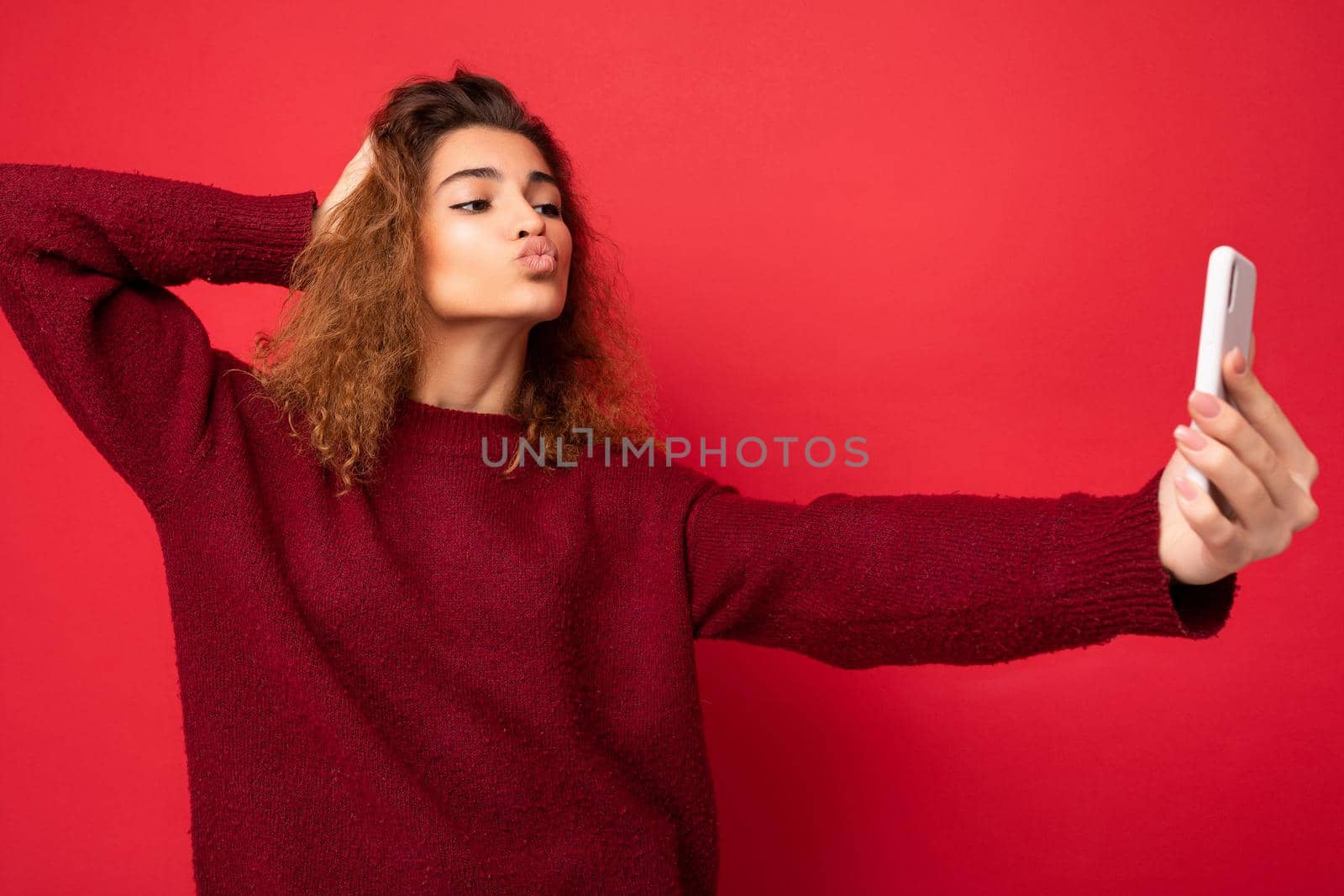 Attractive young blogger woman with curly hair wearing dark red sweater isolated on red background wall holding and using smart phone looking at telephone screen and taking selfie and giving kiss.