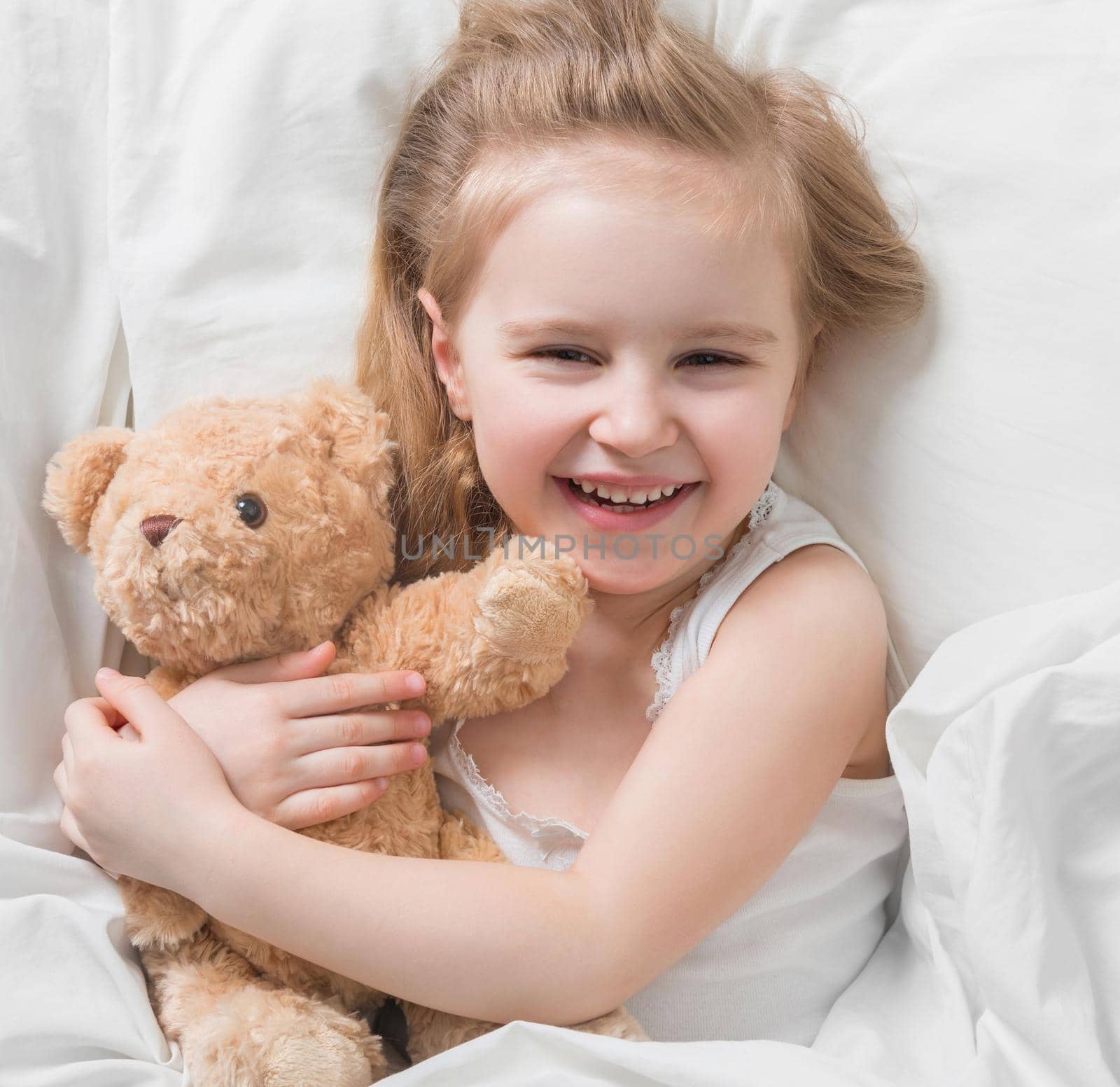 Lovely little blonde girl holding her favorite brown teddy, lying in bed, smiling widely