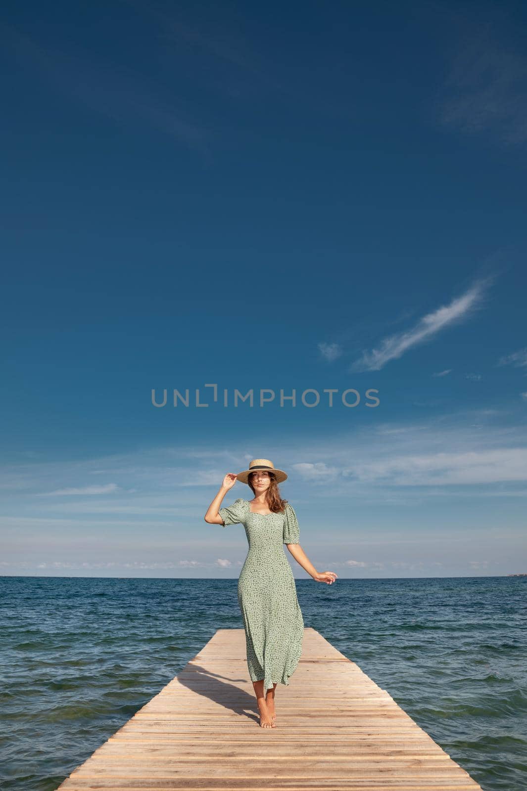 Attractive young caucasian woman in a green dress and a straw hat is walking along a wooden pier against the backdrop of the blue sea and blue sky on a sunny day by yanik88