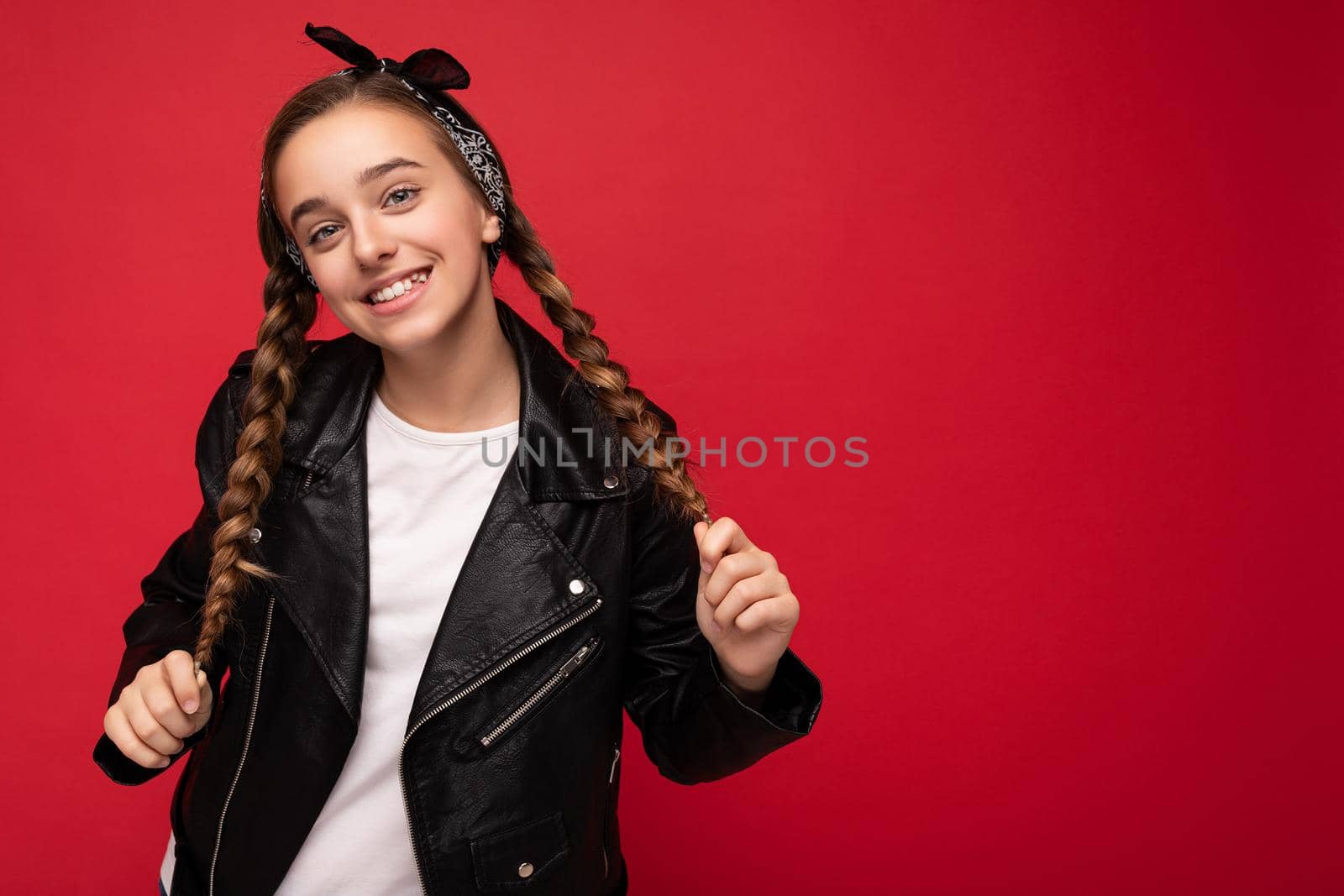 Photo shot of beautiful happy smiling brunette little girl with pigtails wearing trendy black leather jacket and white t-shirt for mockup standing isolated over red background wall looking at camera. Copy space