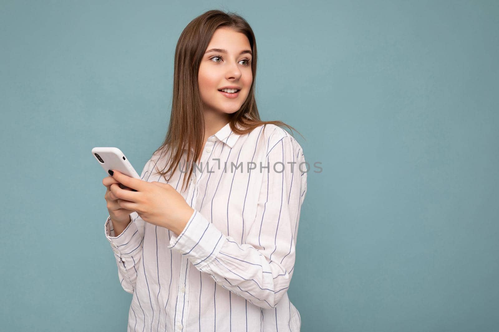 Photo shot of attractive positive good looking young woman wearing casual stylish outfit poising isolated on background with empty space holding in hand and using mobile phone messaging sms looking to the side.