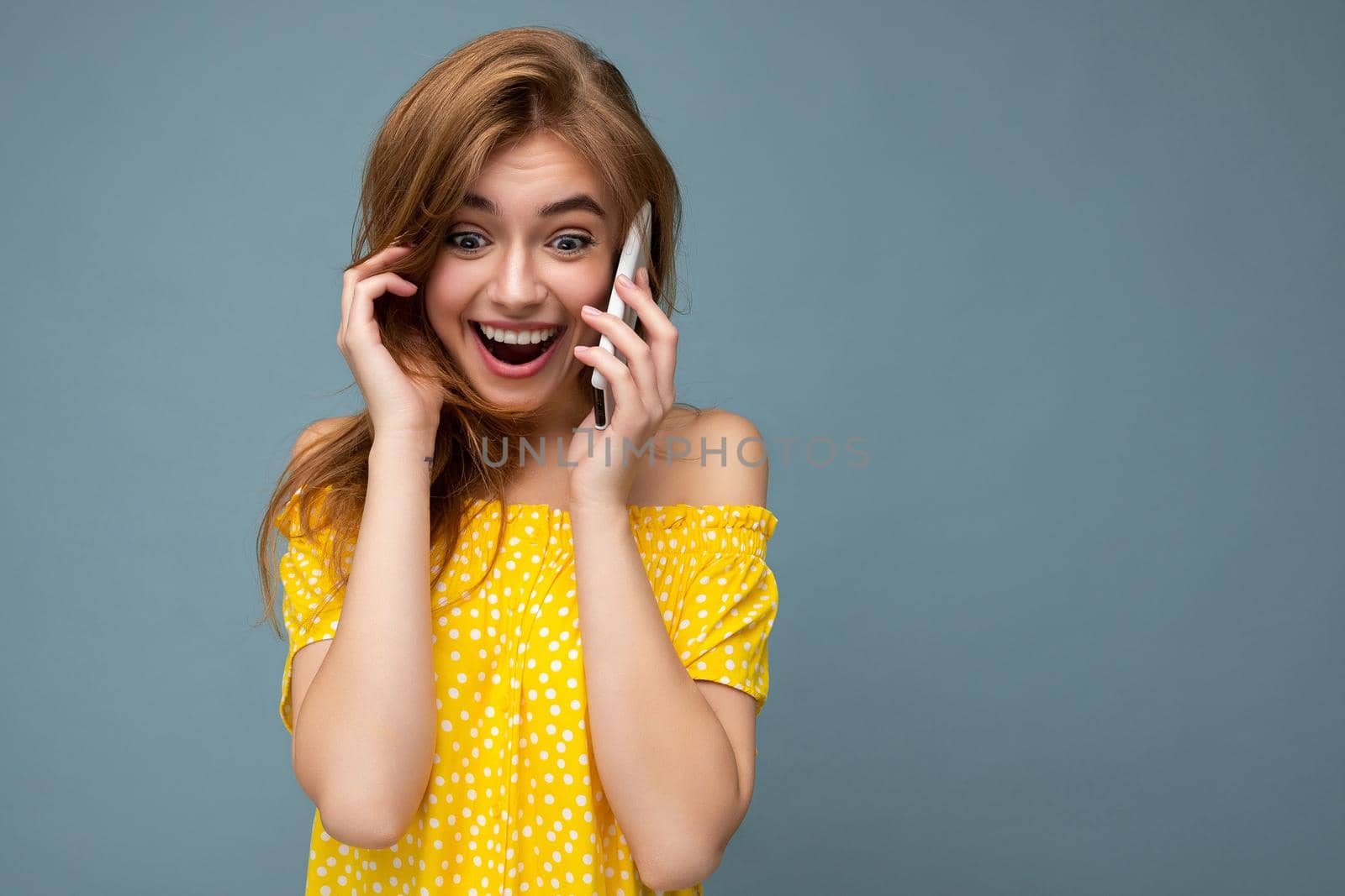 Closeup photo of pretty positive amazed young blonde woman wearing stylish yellow summer dress standing isolated over blue background holding and talking on mobile phone with sincere emotions looking to the side.