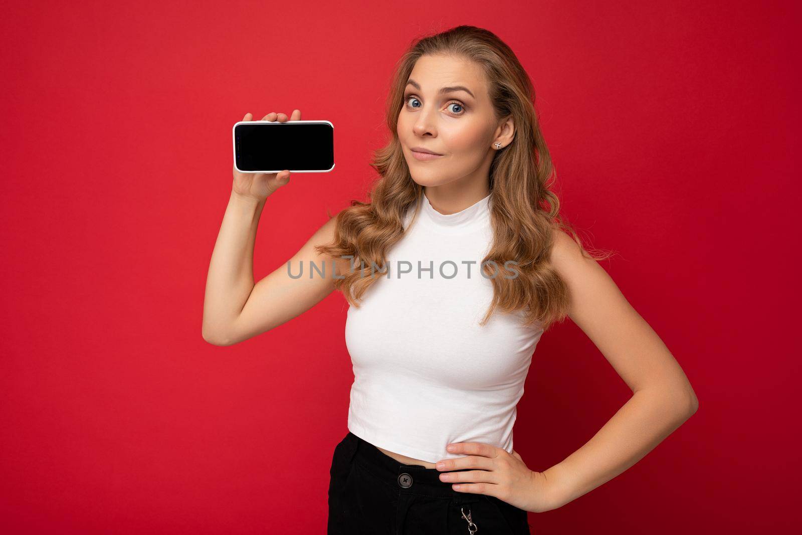 beautiful young blonde woman wearing white t-shirt isolated on red background with copy space holding smartphone showing phone in hand with empty screen for mockup looking at camera.