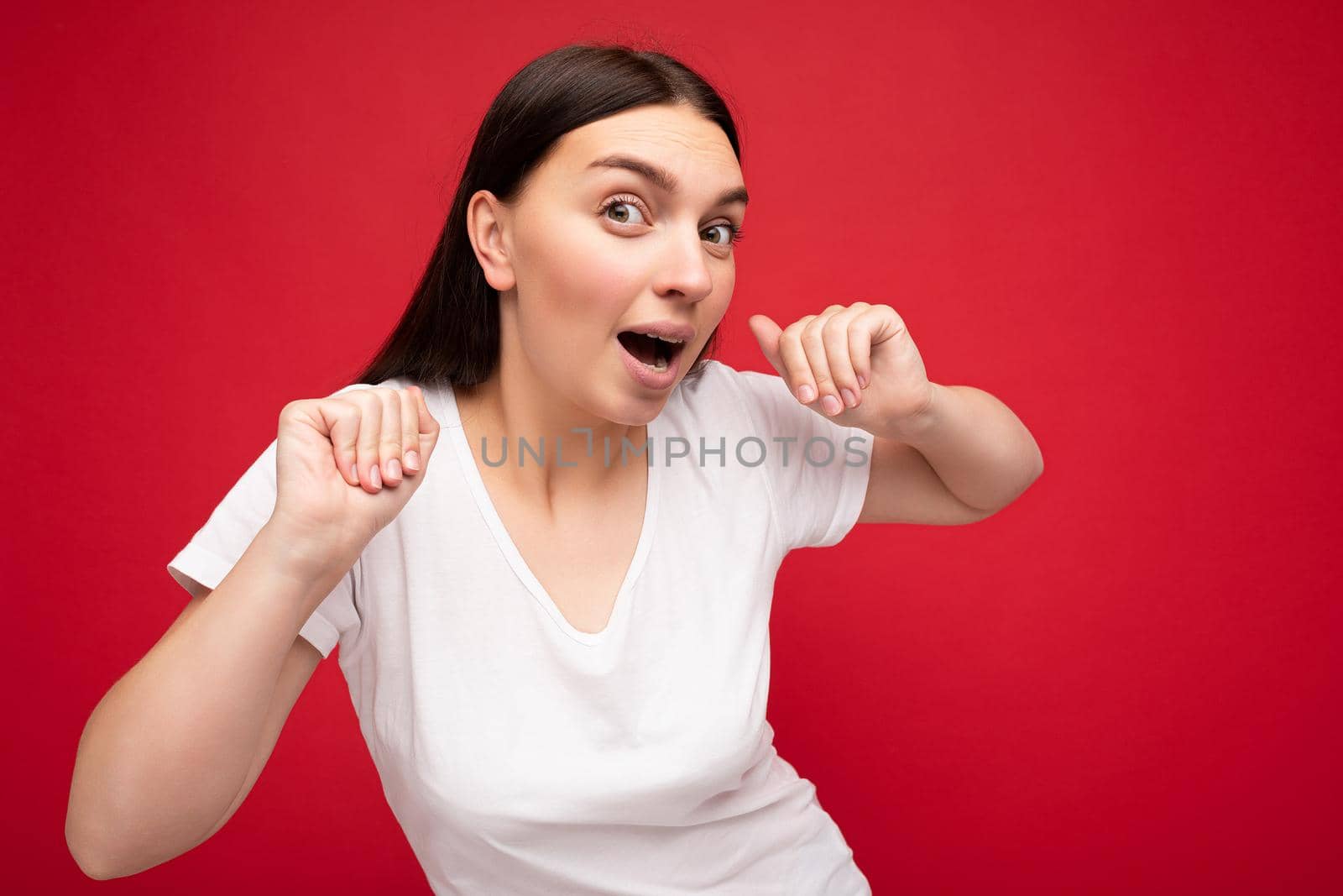 Photo of positive happy surprised young beautiful brunette woman with sincere emotions wearing white t-shirt for mockup isolated over red background with empty space.