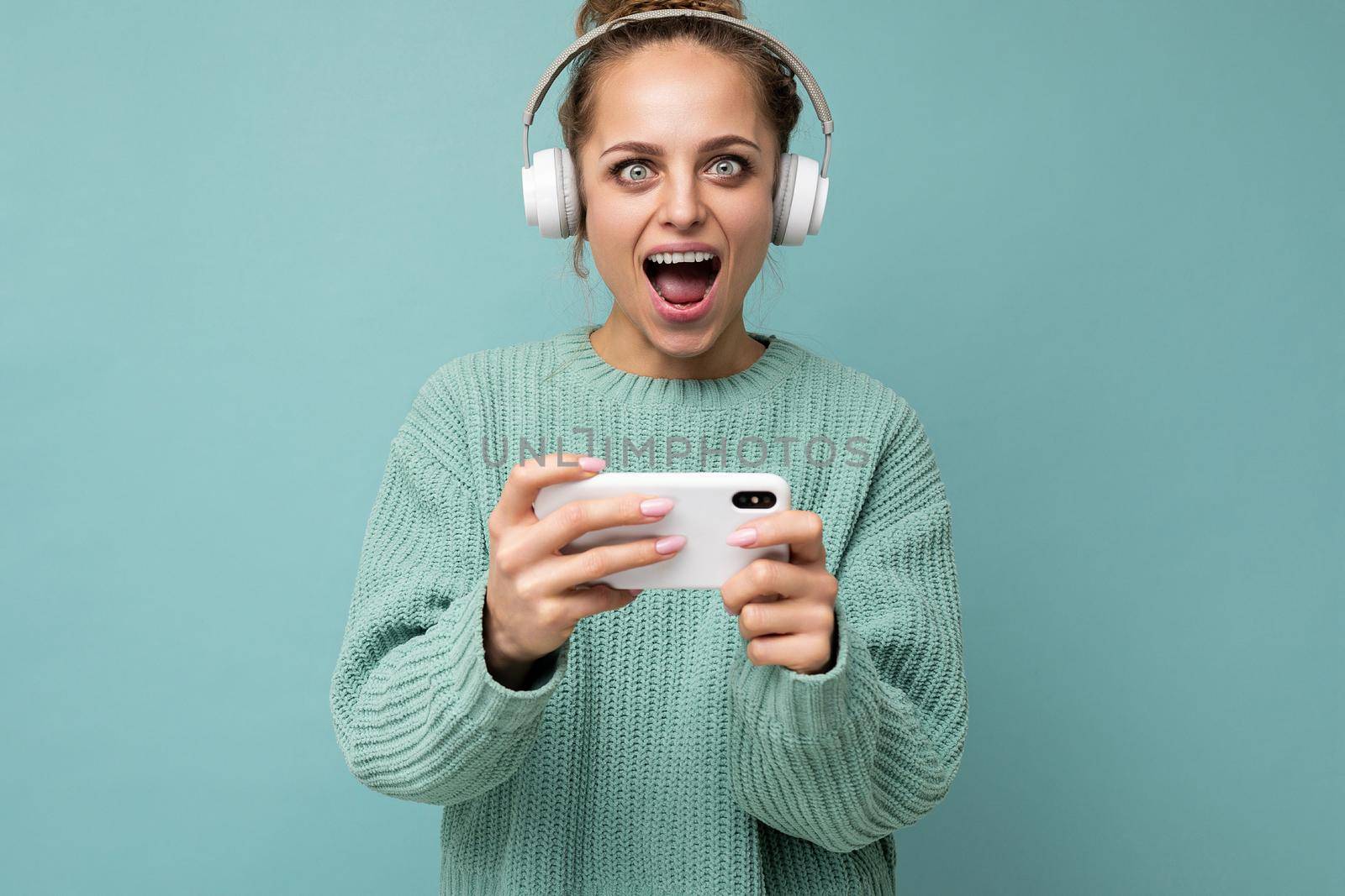 Closeup portrait of attractive emotional positive young woman wearing blue sweater isolated over blue background wearing white bluetooth wireless headphones and listening to music and using mobile phone playing online games looking at camera.