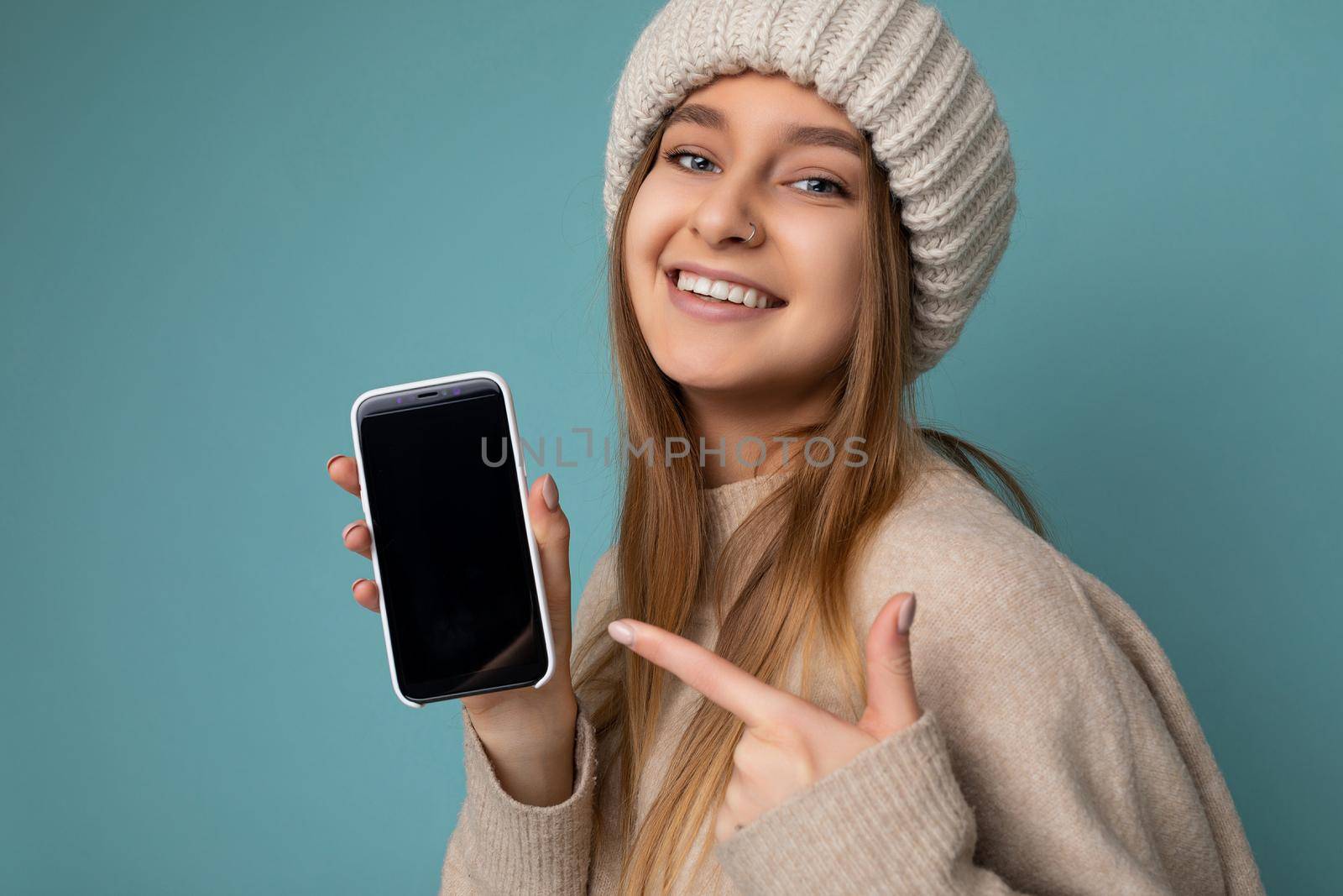 closeup Photo of beautiful smiling young woman good looking wearing casual stylish outfit standing isolated on background with copy space holding smartphone showing phone in hand with empty screen display for mockup pointing at gadjet looking at camera.