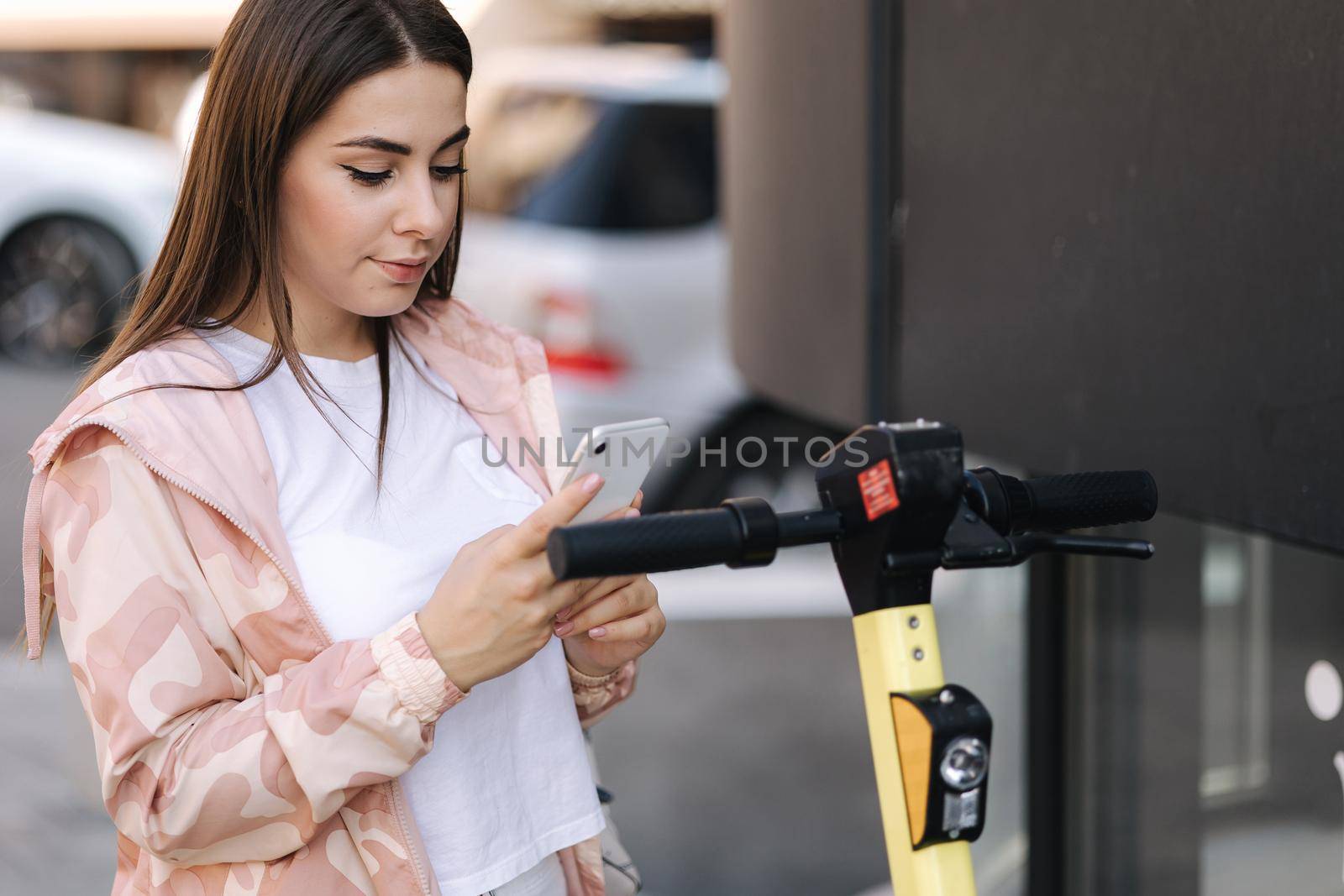 Young woman connect an electric scooter outdoors. Woman using phone for contactless payment. Ecological transportation concept by Gritsiv