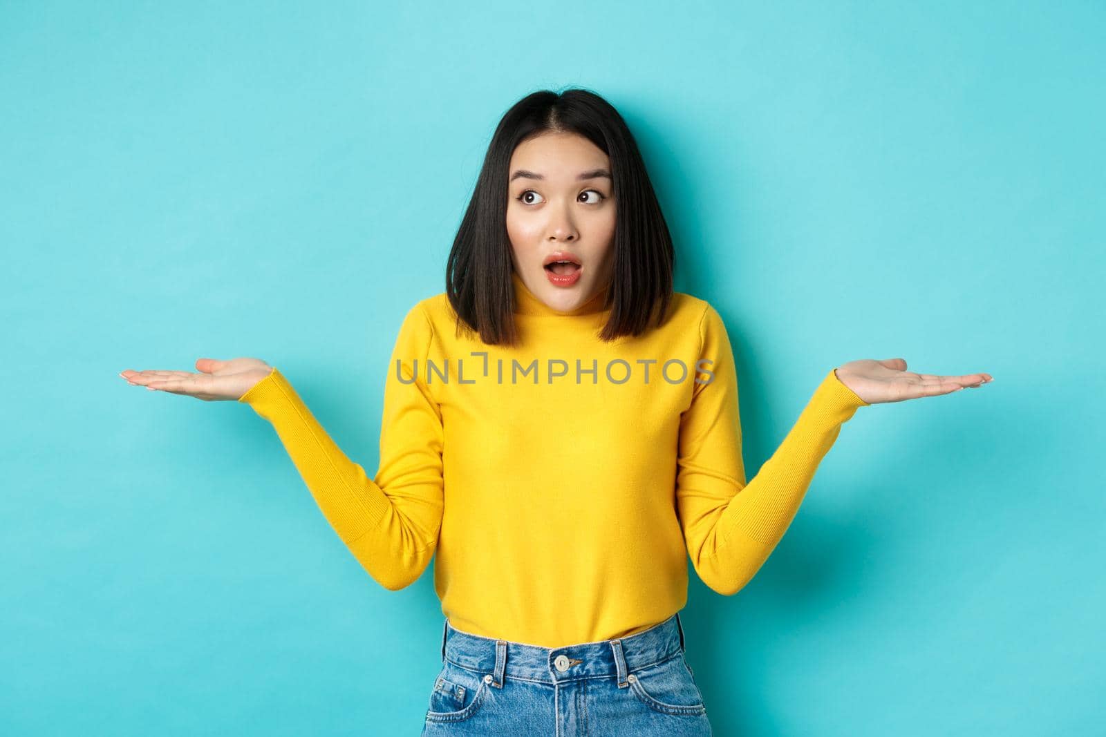 Confused and shocked asian woman dont understand, shrugging with spread hands, gasping and looking left, know nothing, standing over blue background.