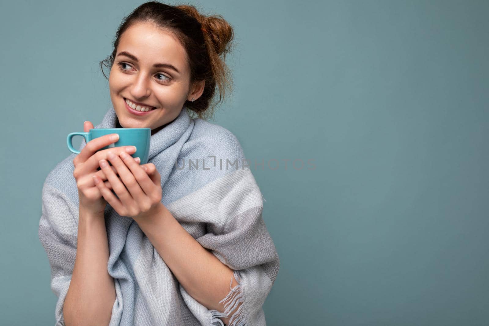 Attractive young happy smiling brunette woman wearing warm blue scarf isolated over blue background holding blue mug drinking coffee and looking to the side. copy space