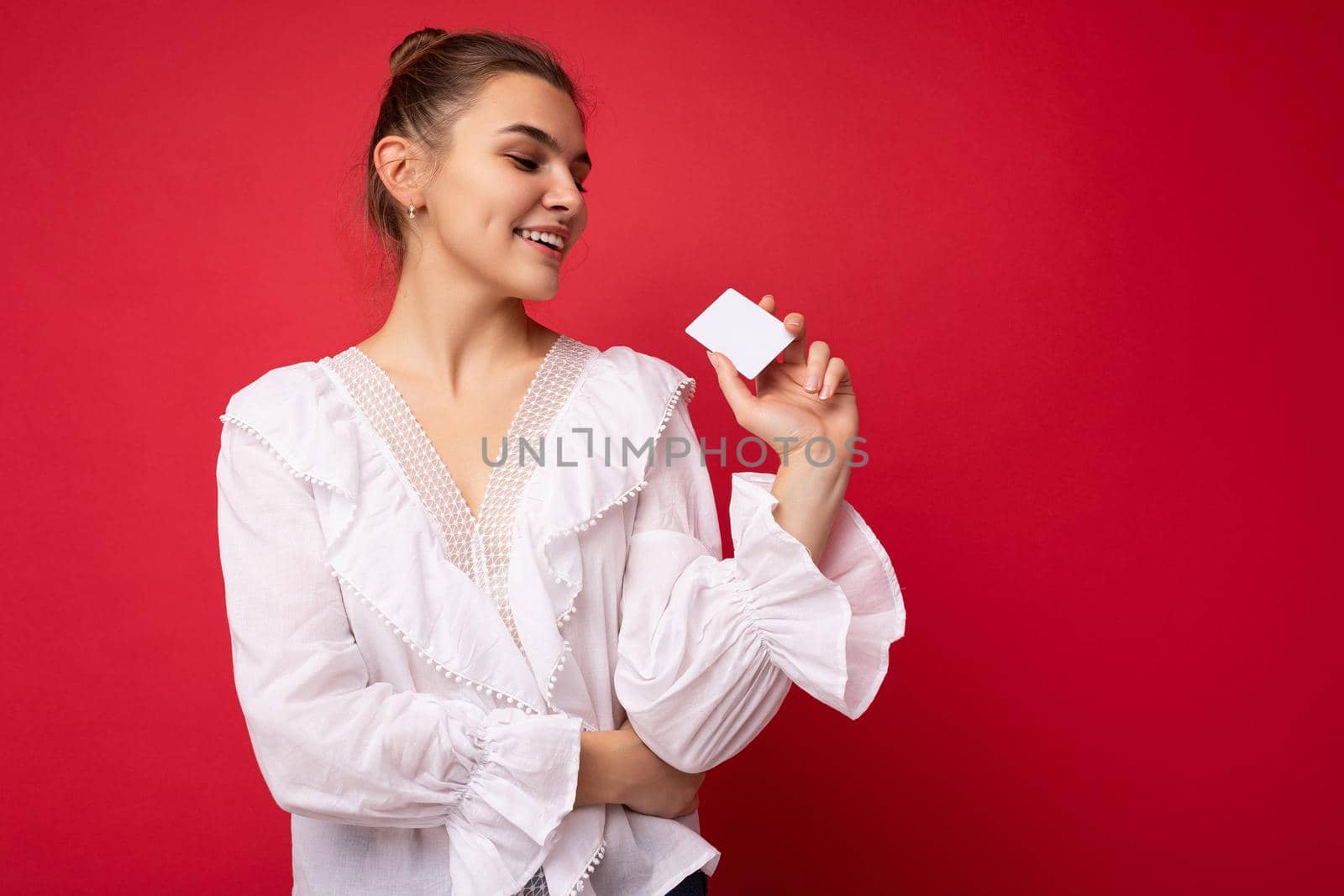 Photo of beautiful happy young dark blonde woman wearing white blouse isolated over red background holding credit card looking at plastic card. empty space