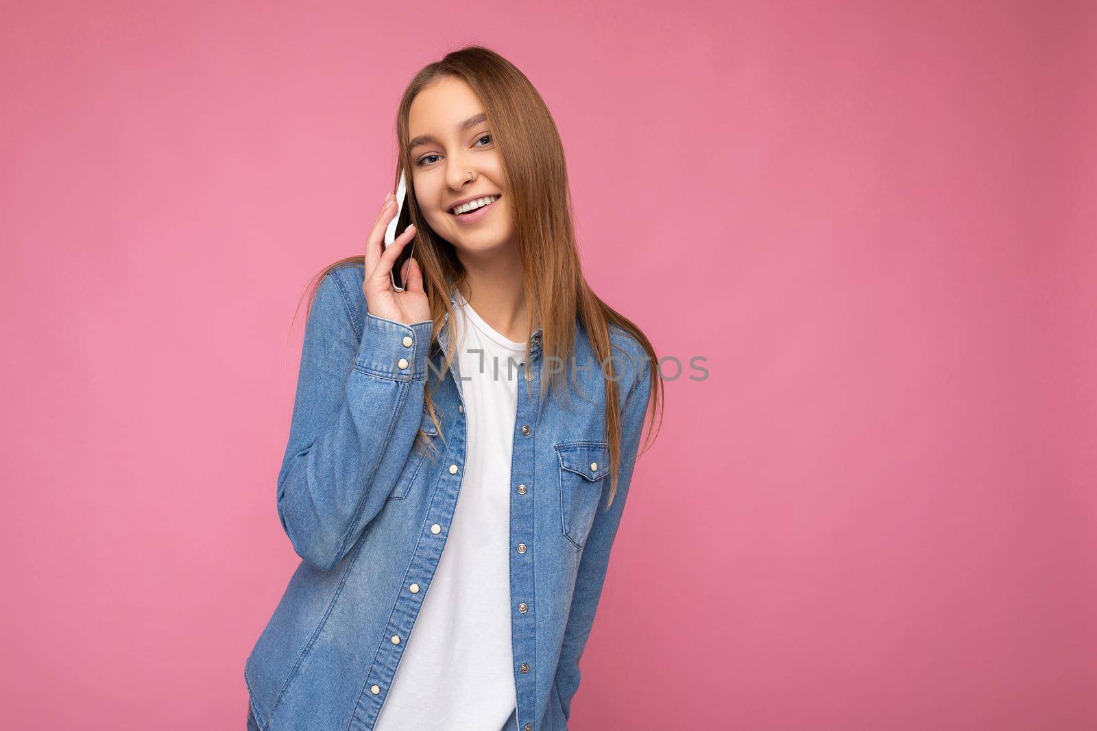 Photo of Attractive sexy positive young blonde woman wearing casual blue jean shirt isolated over pink background holding in hand and talking on mobile phone looking at camera. copy space