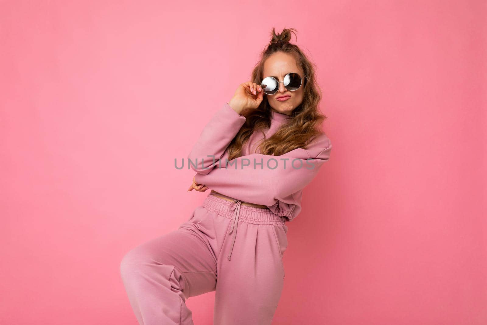 Photo of happy positive cool young beautiful blonde wavy-haired woman with sincere emotions wearing stylish pink sportswear and sunglasses isolated on pink background with copy space and having fun.
