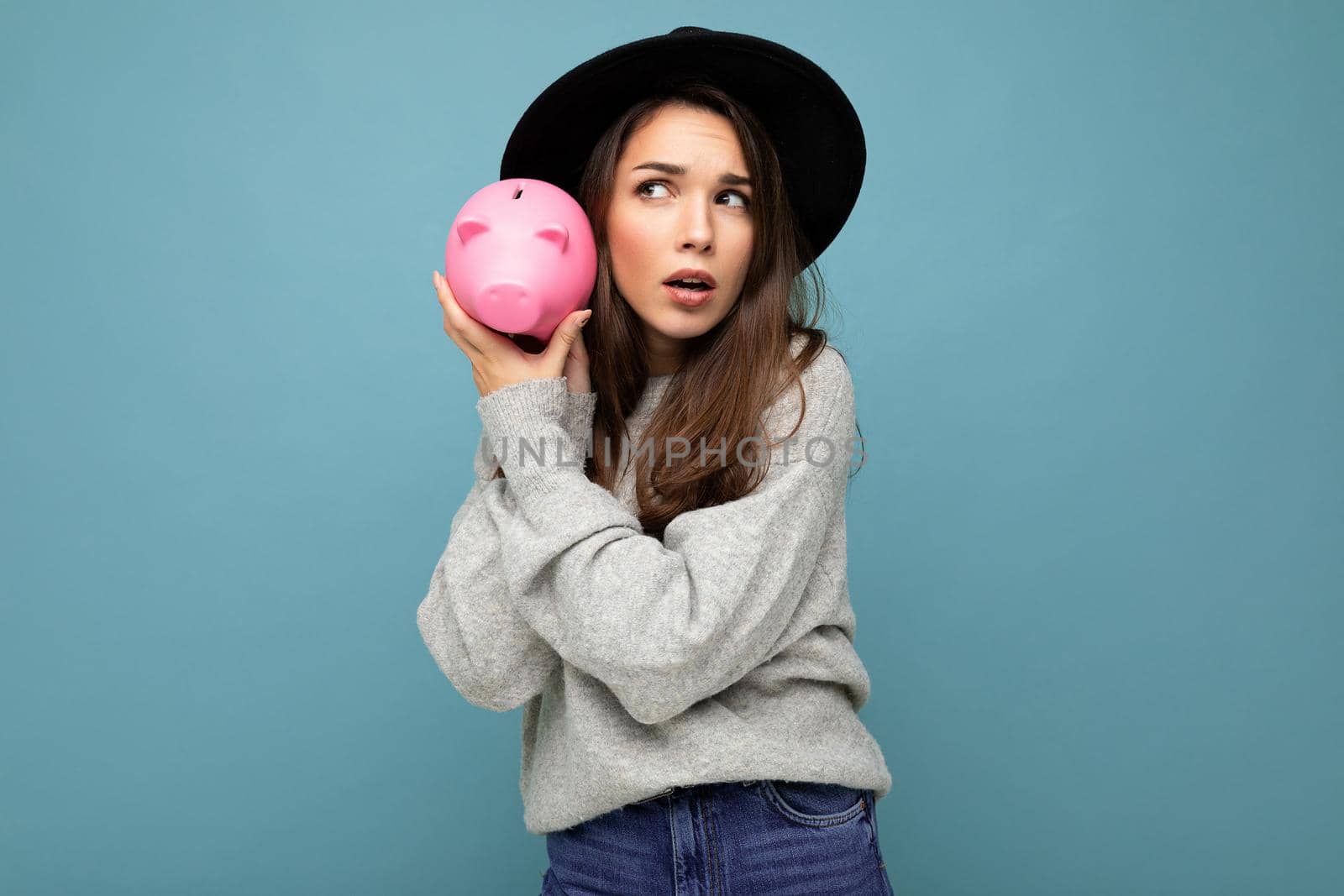 Portrait of positive shocked amazed young beautiful pretty brunette woman wearing gray pullover and black hat isolated over blue background with free space and holding pink piggy box. Money box concept.