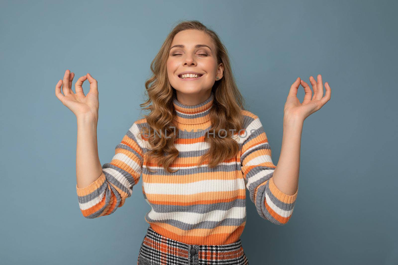 Portrait of smiling happy young attractive blonde curly woman with sincere emotions wearing trendy striped pullover isolated on blue background with copy space and doing mediation gesture with fingers. Yoga concept.