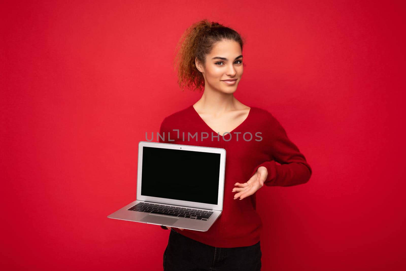 side profile portrait photo of beautiful charming fascinating young curly dark blond lady wearing red sweater standing isolated over red wall background holding computer laptop with empty copy space mock up pointing to the netbook monitor and keyboard looking at camera and thinking about something with sincere expressions.