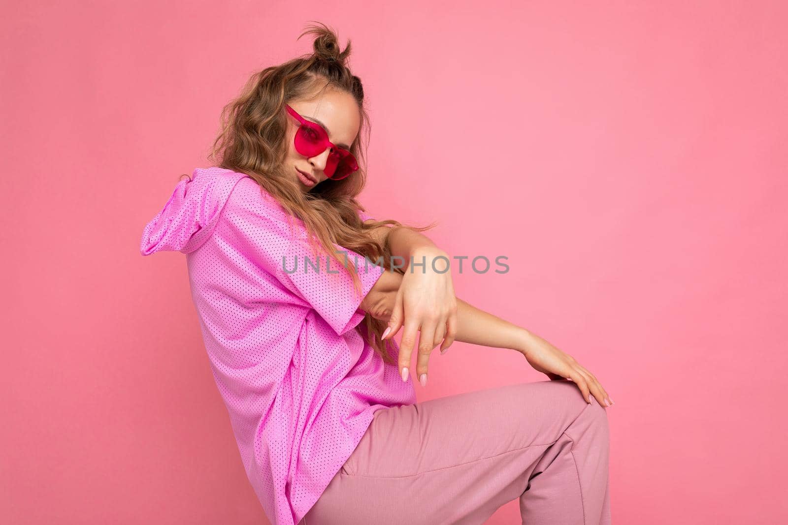 Charming positive young blonde curly woman isolated on pink background wall wearing casual pink t-shirt and stylish colourful sunglasses looking at camera and dancing.