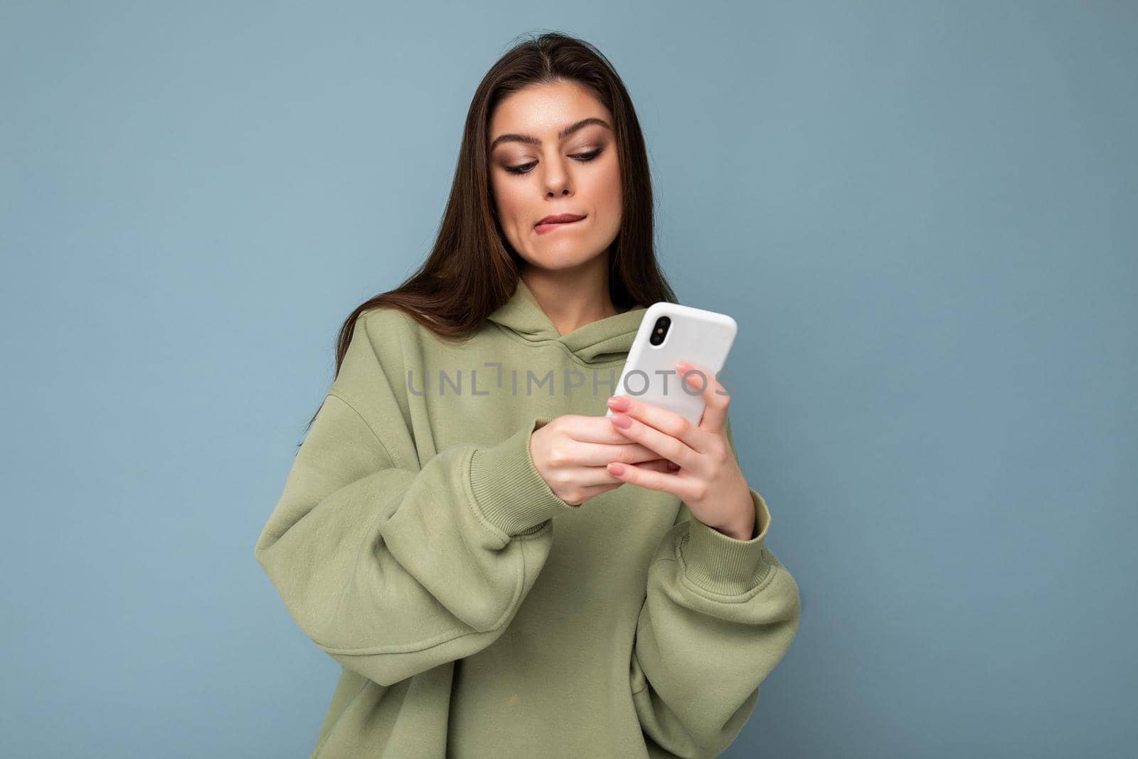 Beautiful young brunette woman wearing stylish green hoodie using mobile phone writing sms isolated on background looking at smartphone screen.