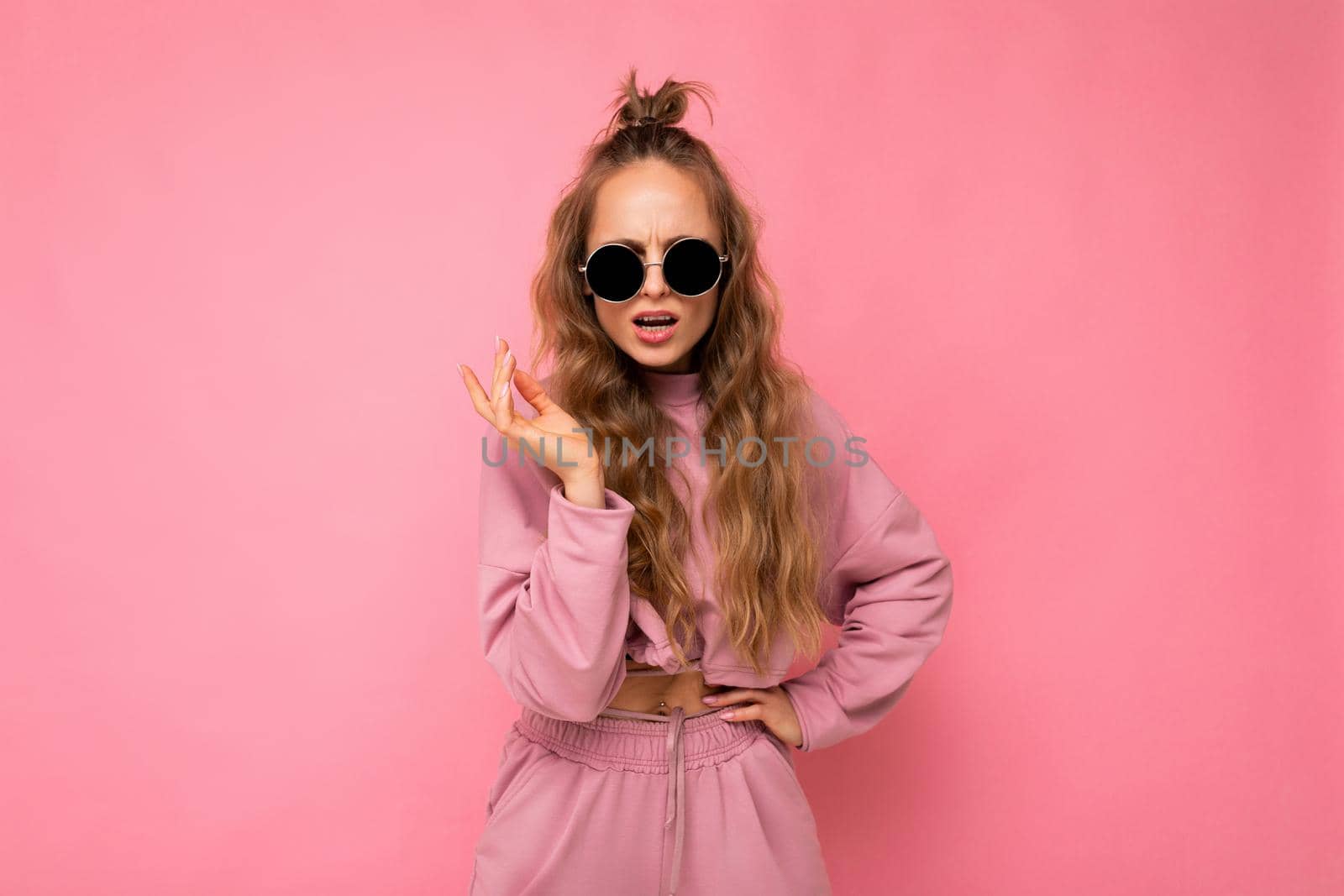 Fascinating dissatisfied adult blonde curly woman isolated over pink background wall wearing casual pink sport clothes and stylish sunglasses looking at camera.