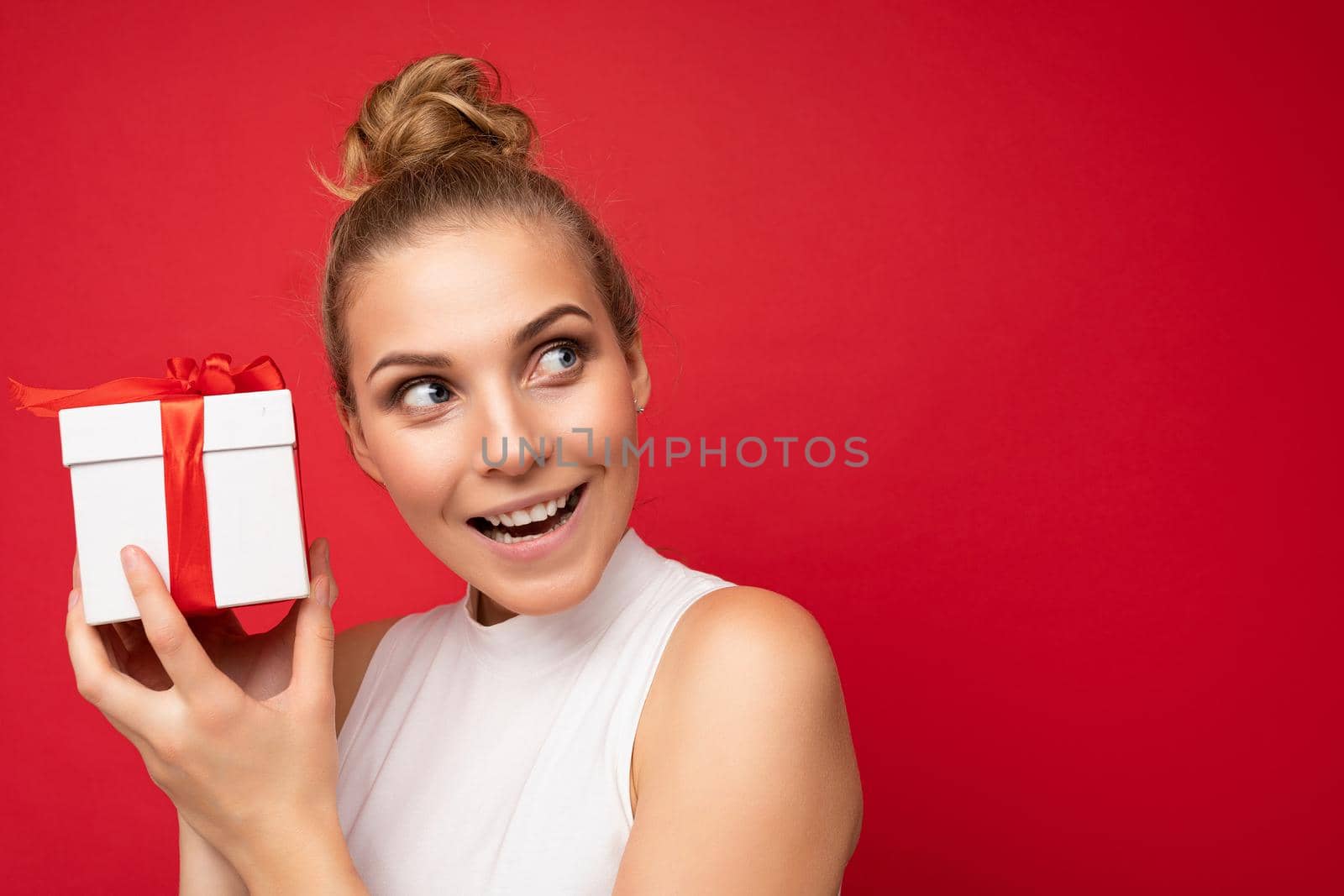 Beautiful happy young blonde woman isolated over colourful background wall wearing stylish casual clothes holding gift box and looking to the side. Copy space, mockup