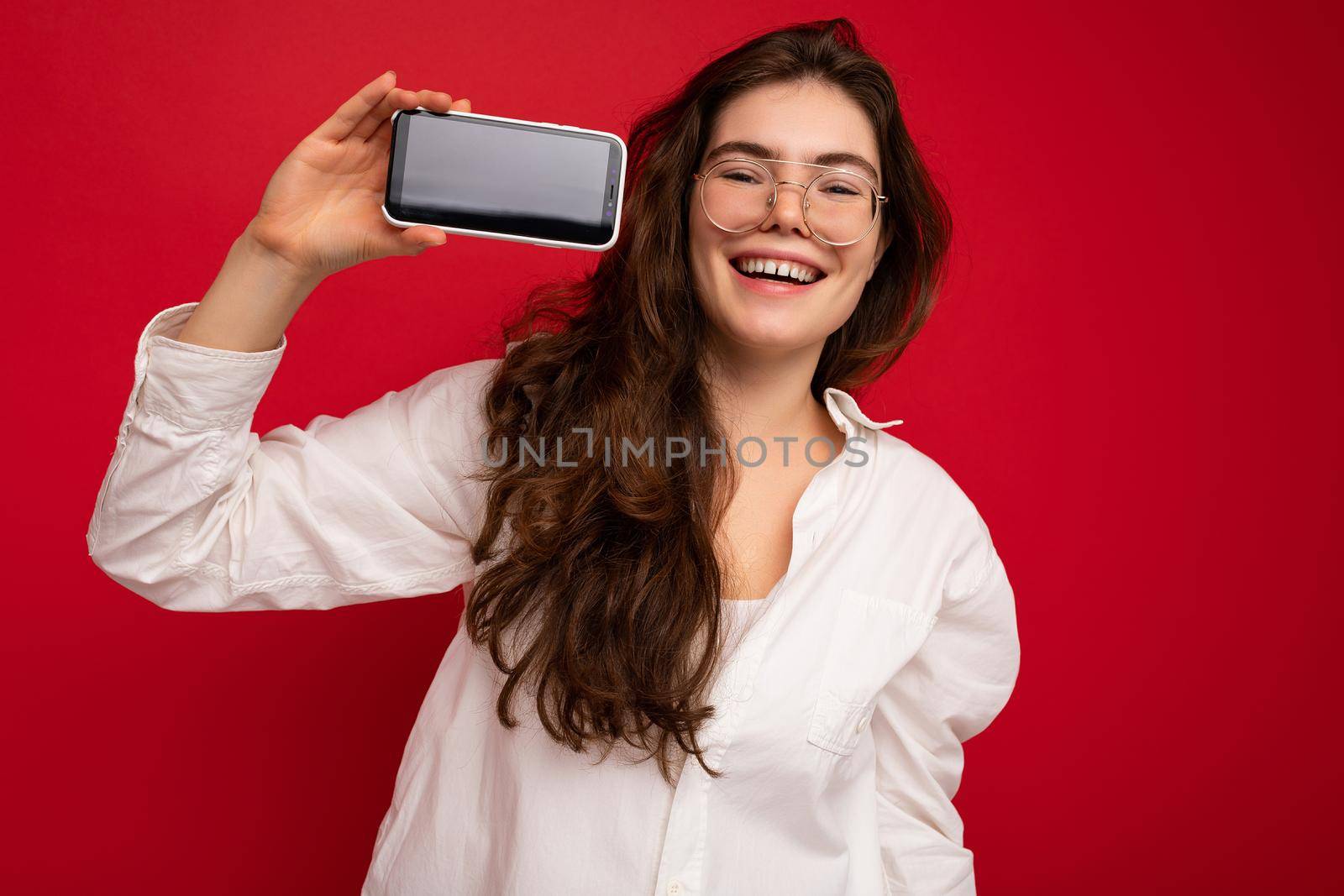 Photo of beautiful smiling young woman good looking wearing casual stylish outfit standing isolated on background with copy space holding smartphone showing phone in hand with empty screen display for mockup looking at camera.