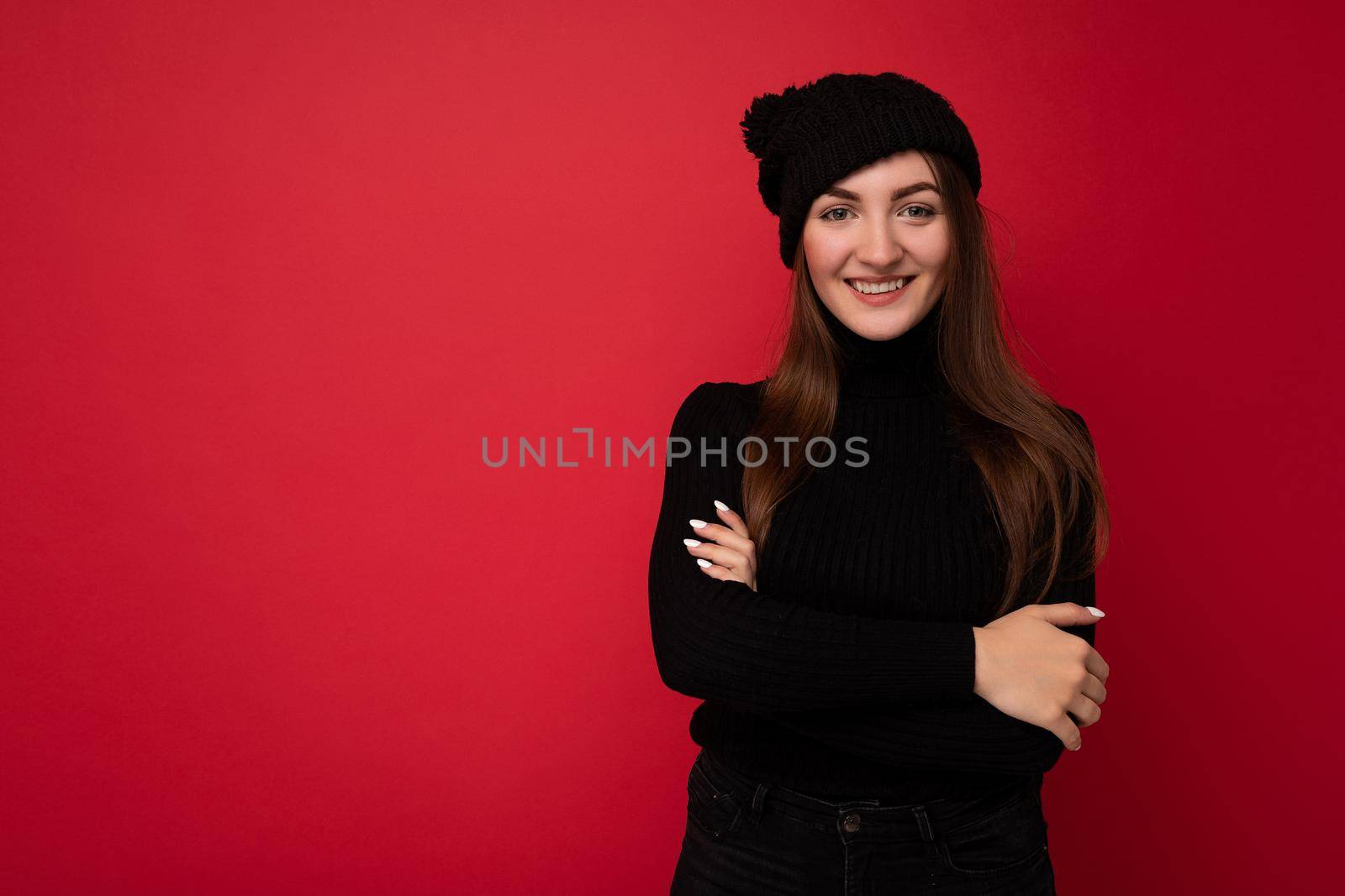 Beautiful positive young brunette woman wearing black sweater and black hat standing isolated over red background looking at camera and smiling keeping hands crossing.