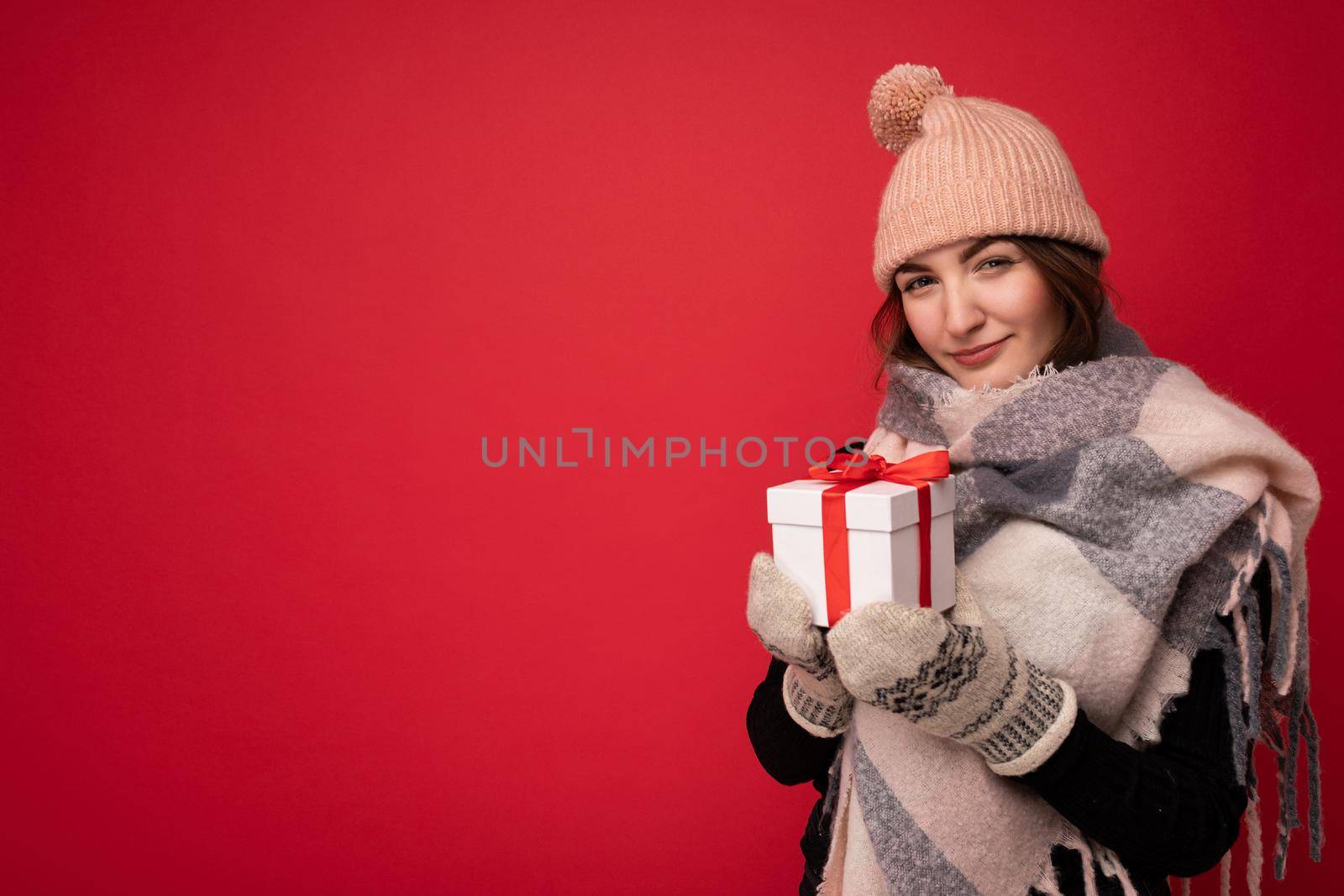 Cozy shot of charming happy cute young brunette woman isolated over red background wall wearing winter scarf hat and mittens holding white gift box with red ribbon and looking at camera. Empty space
