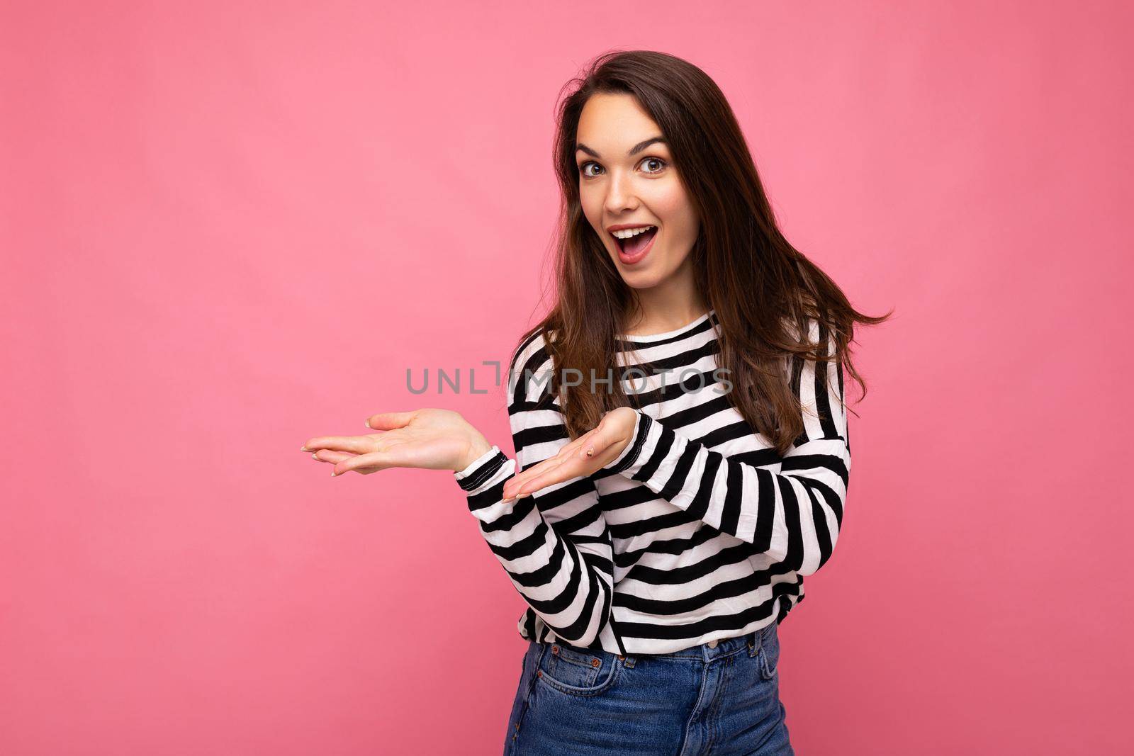 Portrait photo of young happy positive beautiful brunette woman with sincere emotions wearing casual striped pullover isolated on pink background with copy space and pointing hands at free space for text.