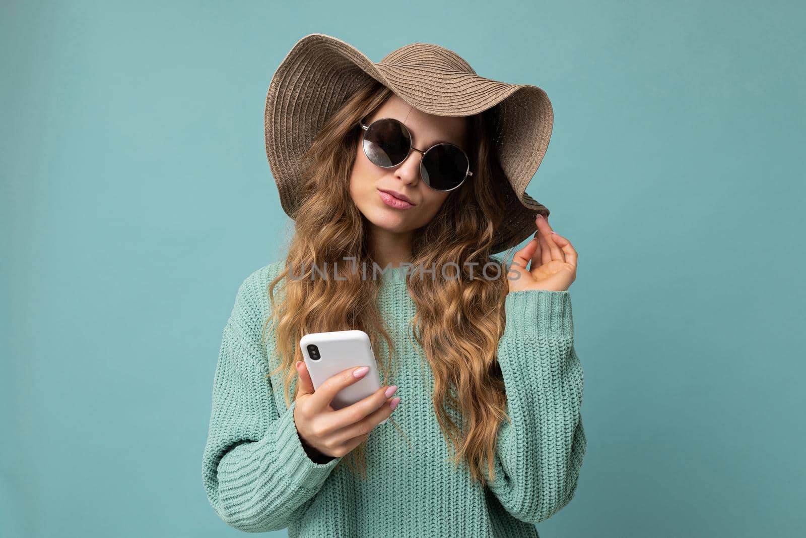 Attractive young blonde woman wearing blue sweater hat and sunglasses standing isolated over blue background surfing on the internet via phone looking at gadjet.