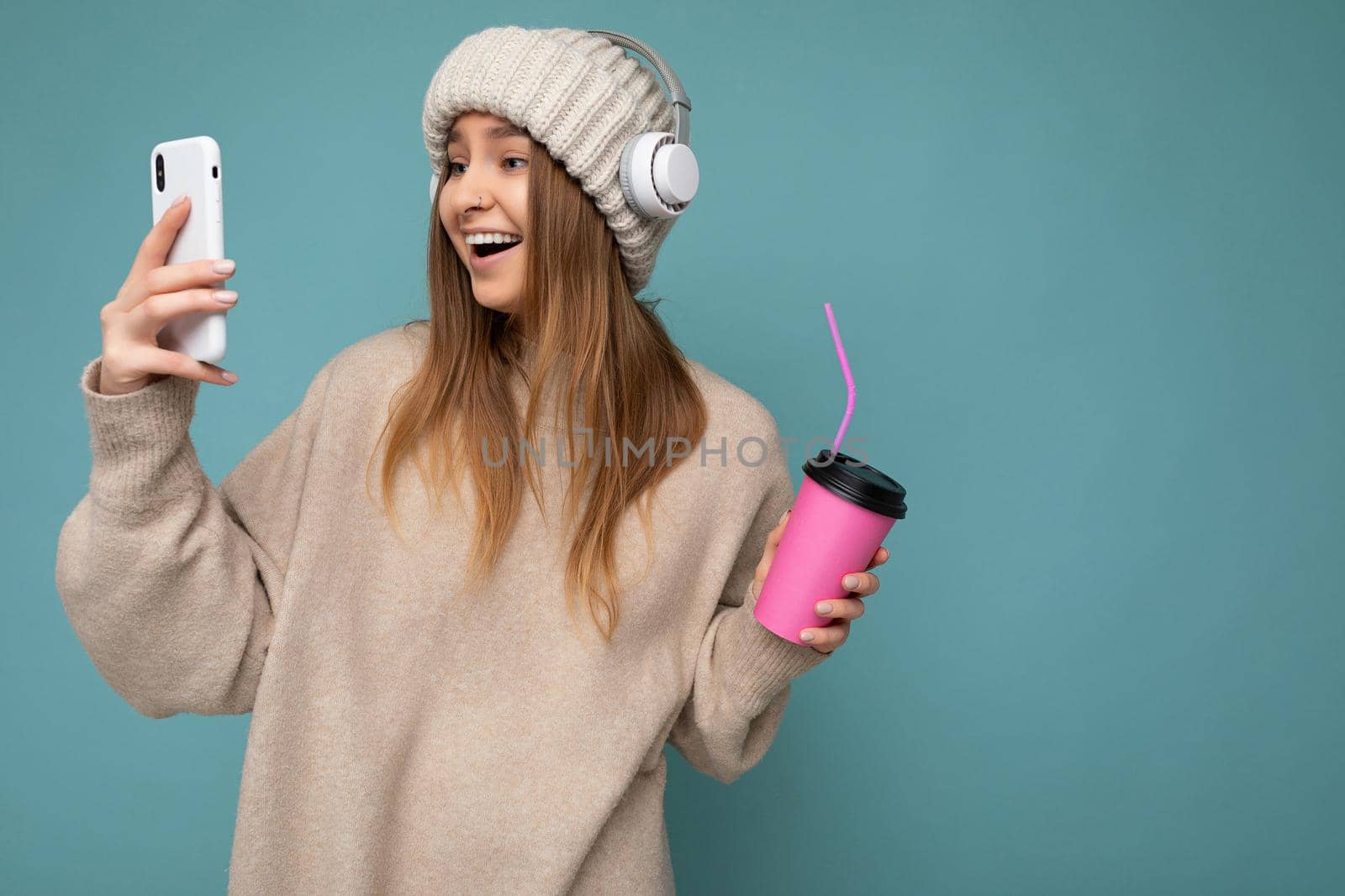 Attractive overjoyed delightful young blonde woman wearing beige sweater and beige hat white headphones isolated over blue background holding in hand and using mobile phone drinking beverage and listening to music looking at gadjet display. copy spcae