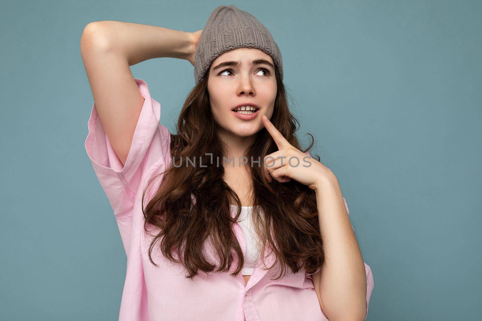 Photo shot of attractive thoughtful young brunette female person standing isolated over blue background wall wearing pink shirt and gray hat looking to the side.