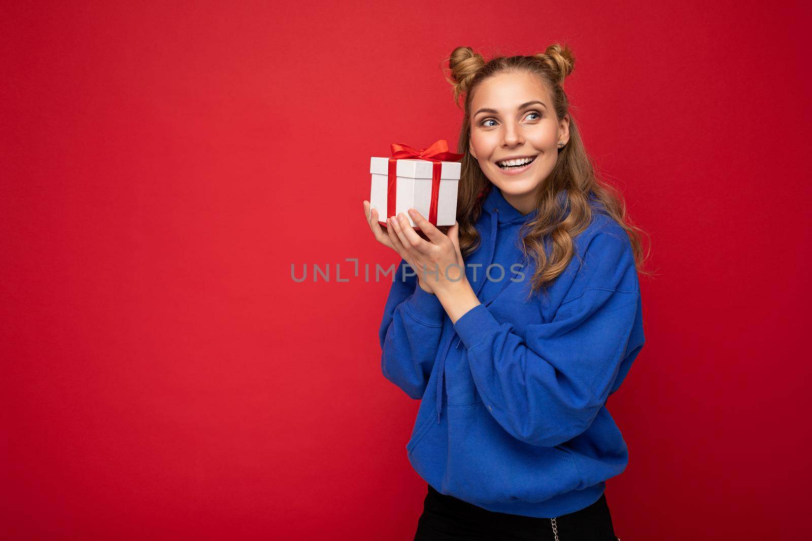 Shot of attractive happy smiling young blonde woman isolated over red background wall wearing blue trendy hoodie holding gift box and looking to the side. Copy space, mockup
