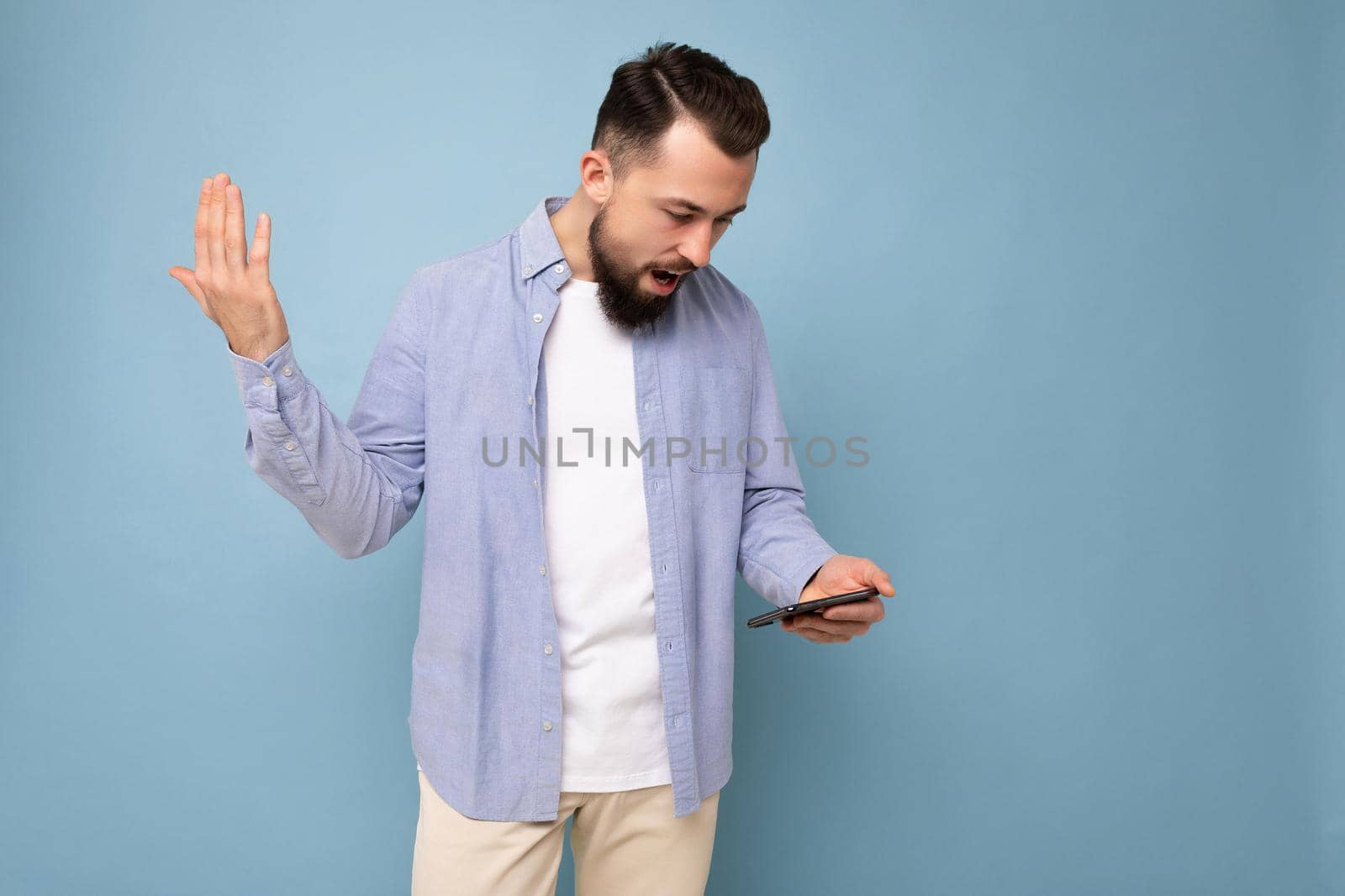 Photo shot of dissatisfied handsome bearded good looking young man wearing casual stylish outfit poising isolated on background with empty space holding in hand and using mobile phone messaging sms looking at smartphone display screen.