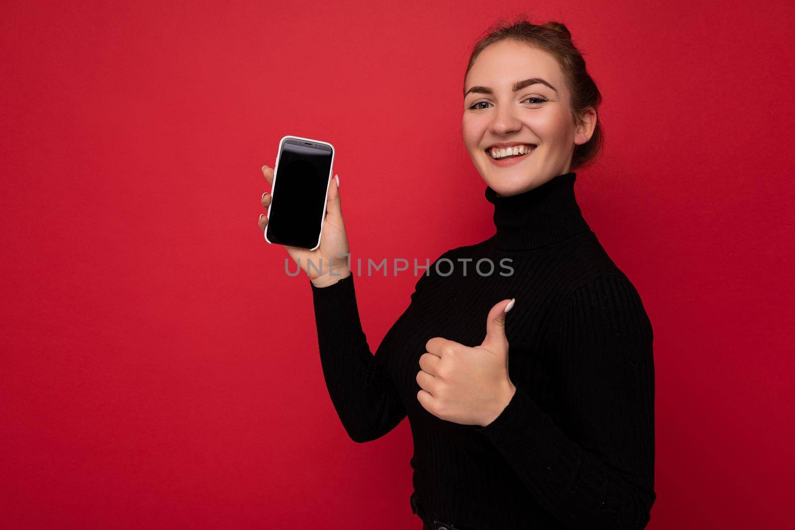 side-profile Photo of attractive positive young brunette woman wearing black sweater standing isolated over red background showing mobile phone with empty screen for mockup looking at camera and showing thumbs up gesture by TRMK