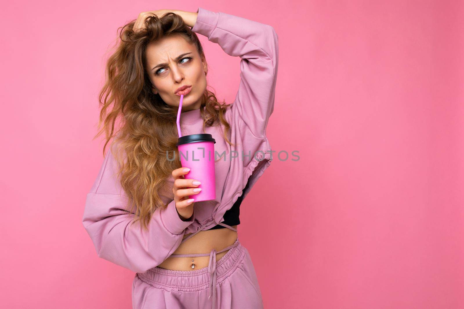 Photo shot of beautiful sad upset young blonde woman wearing pink sport clothes isolated over pink background holding paper coffee cup for mockup drinking and looking to the side by TRMK