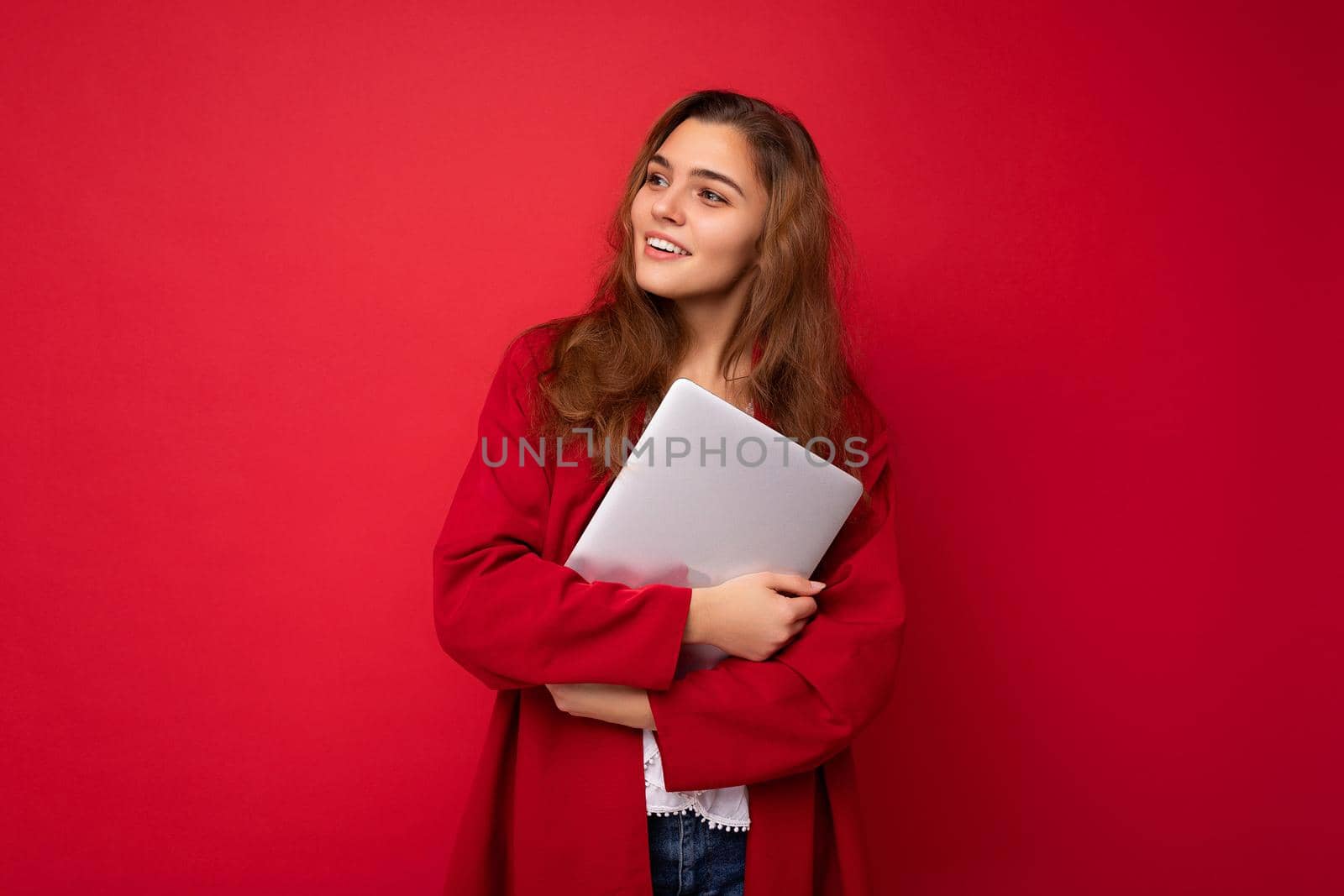 shot of mysterious Beautiful young dark hair curly woman holding close laptop wearing red cardigan and white blouse looking to the side isolated over red background.