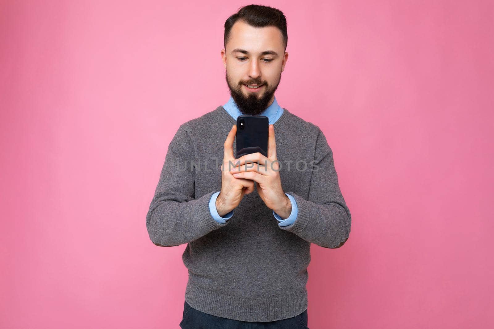 handsome good looking brunet bearded young man wearing grey sweater and blue shirt isolated on pink background with empty space holding in hand and using mobile phone communicating online looking at device screen.
