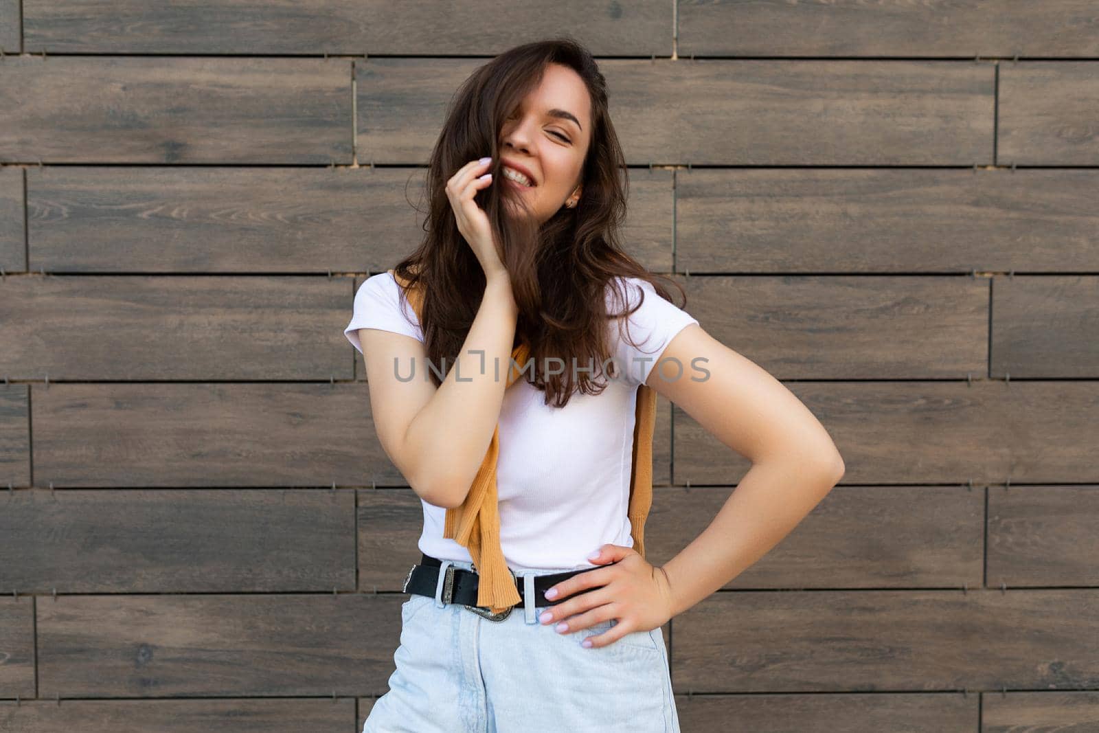 Portrait of successful smiling joyful happy young brunet woman wearing casual white t-shirt and jeans with yellow sweater poising near brown wall in the street and having fun by TRMK