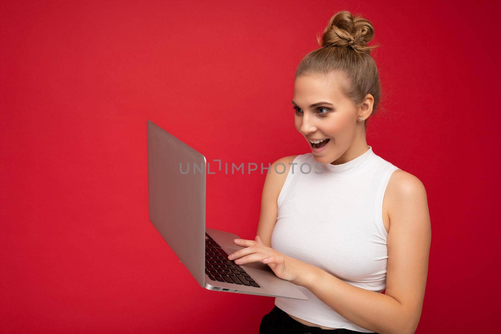 Side profile photo shot of beautiful happy amazed surprised overjoyed joyful blond young woman with gathered hair wearing white t-shirt using computer laptop typing on keyboard looking at netbook monitor screen isolated over red wall background by TRMK