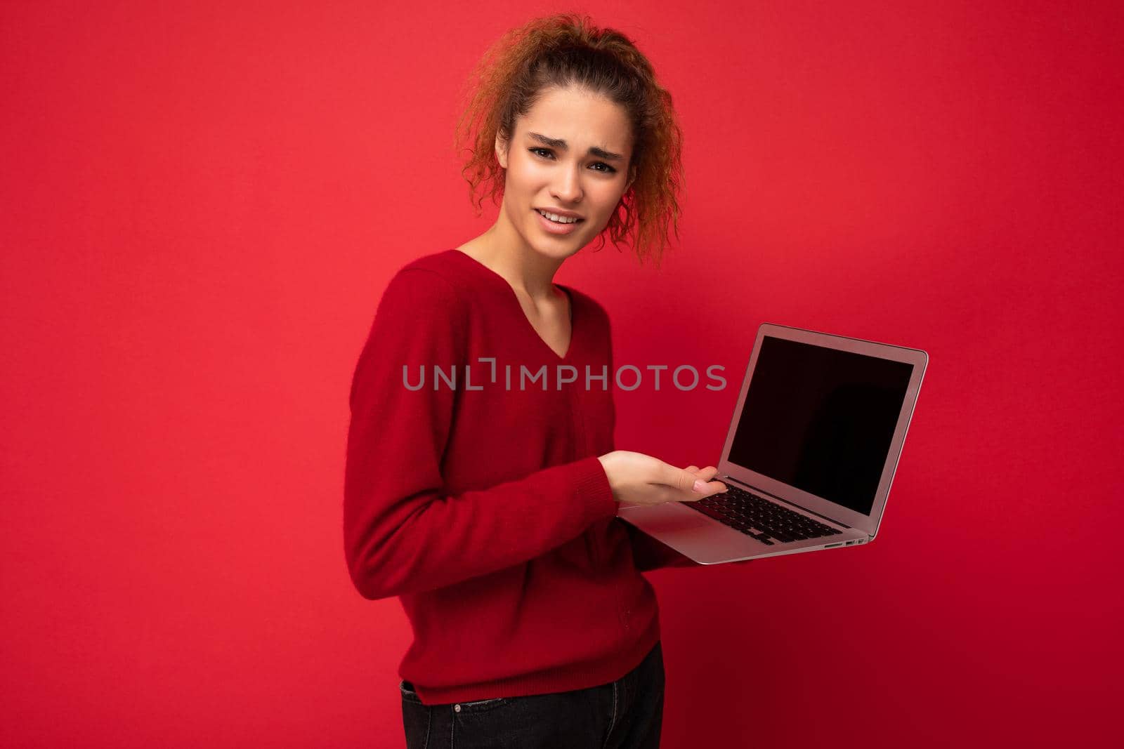 Close-up portrait of beautiful amazed surprised astonished dark blond woman holding laptop computer looking at camera typing on keyboard wearing red sweater isolated over red wall background by TRMK